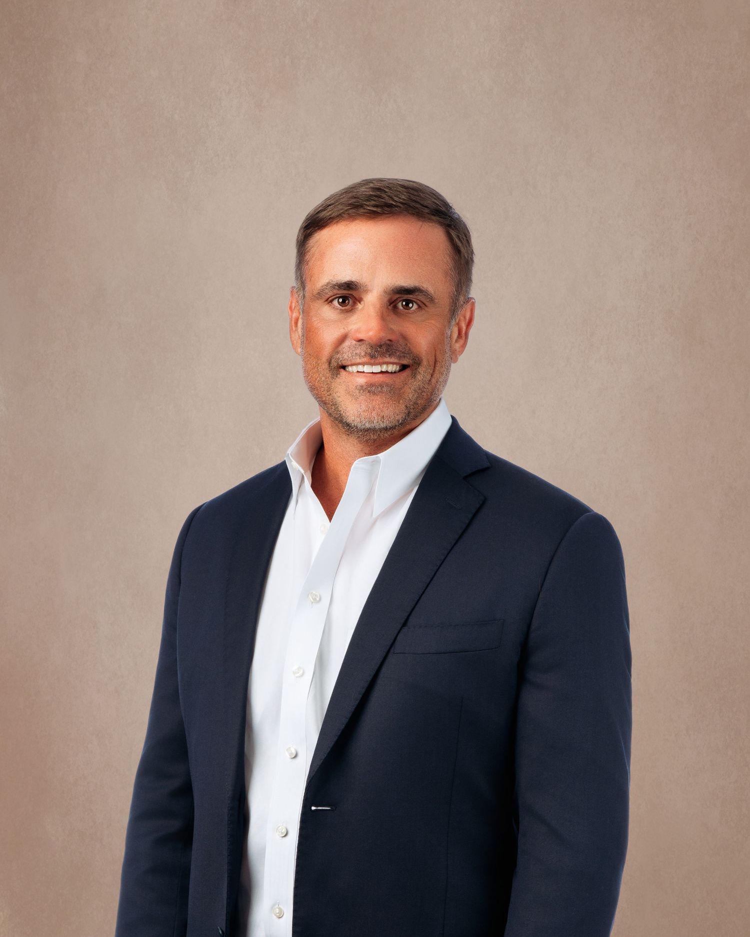 Professional headshot of a smiling middle-aged man in a navy blazer and white shirt against a neutral background.