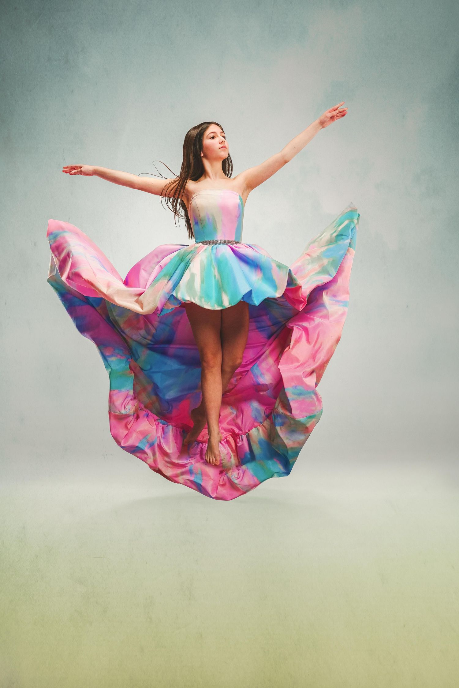 A young woman leaps joyfully in a flowing multicolored pink and blue ball gown dress against a soft studio background.
