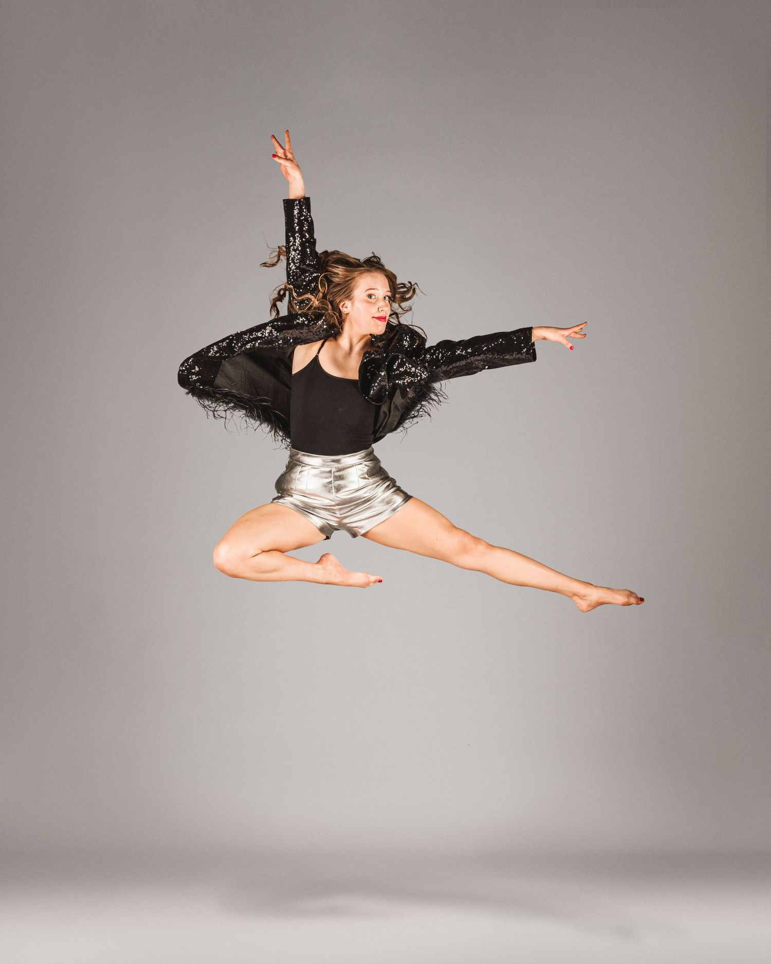 A dancer leaps gracefully in silver shorts and black sequined jacket against a gray studio background.