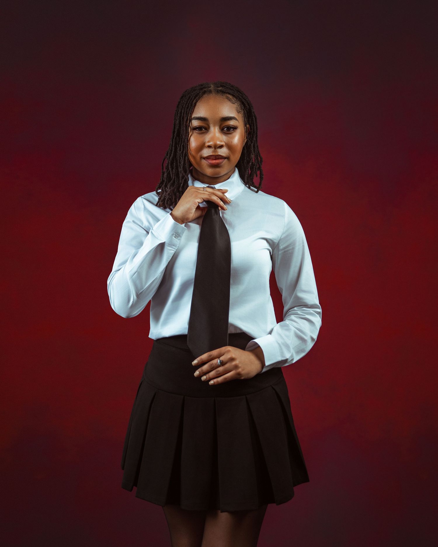 Young Black woman in white shirt, black tie and pleated skirt poses confidently against dark red background.