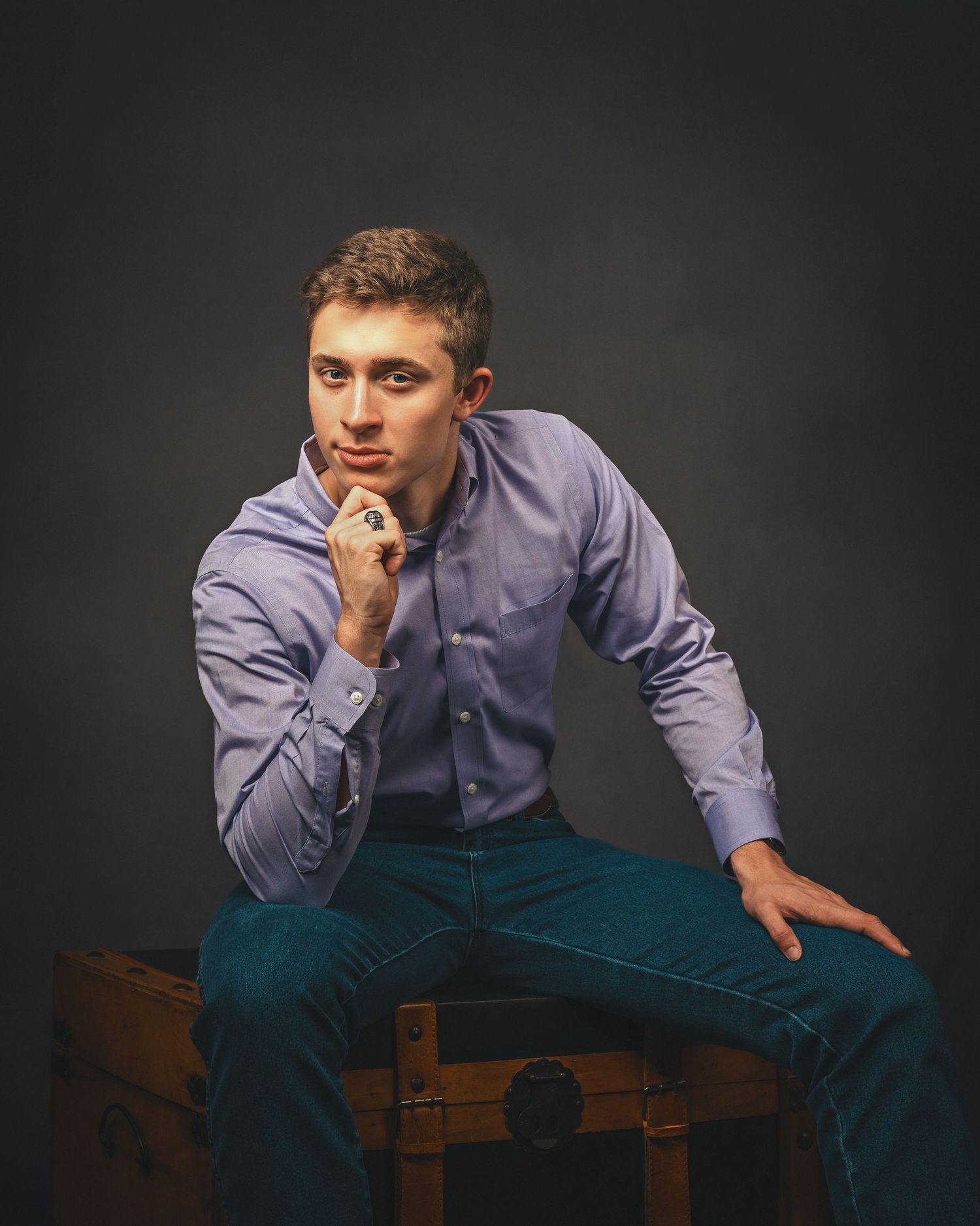 Young man in blue shirt and jeans sits thoughtfully on vintage trunk against dark studio background.
