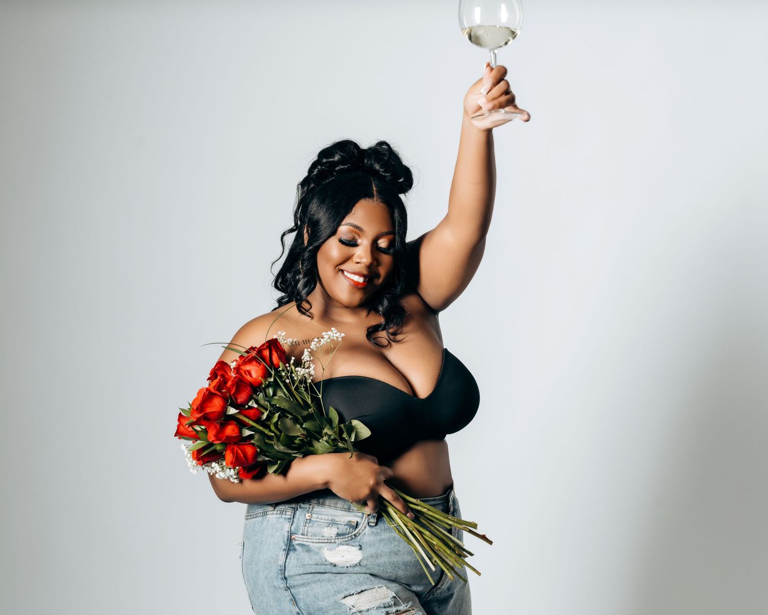 A plus-size woman in a black bralette and jeans holds red roses and raises a wine glass in a studio.