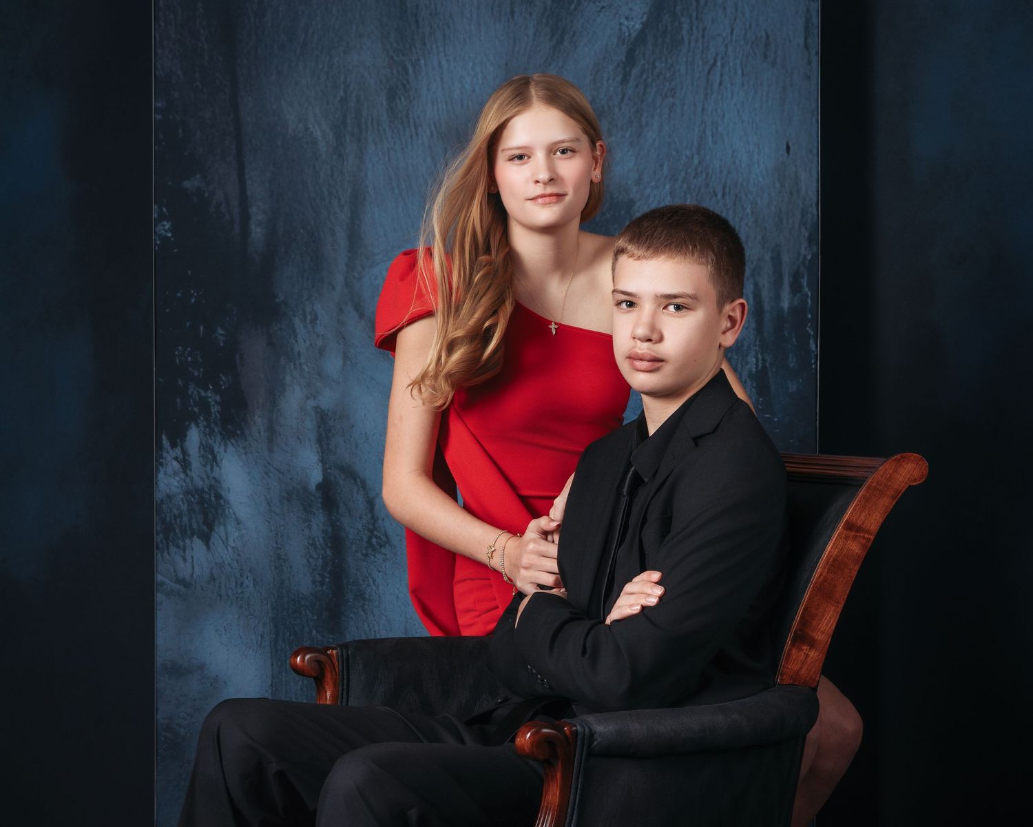 Two siblings pose for a studio portrait, girl in red dress standing behind boy in black suit seated in ornate chair.