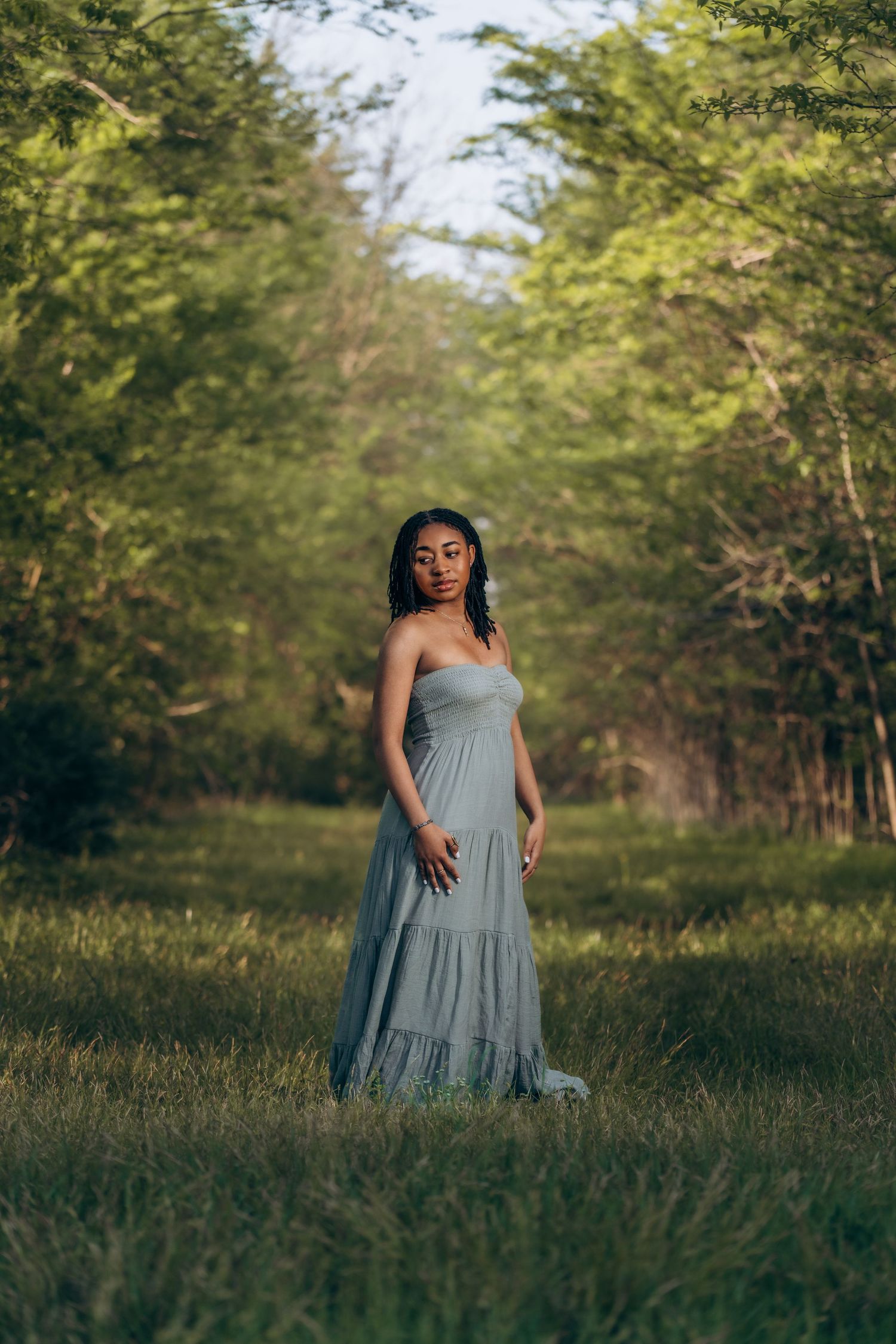 A young Black woman in a flowing gray strapless maxi dress stands gracefully in a lush green forest clearing.