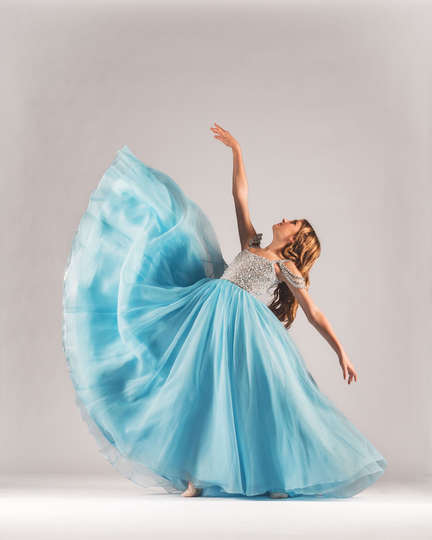 A dancer in a flowing turquoise ball gown gracefully poses with arms extended in a professional studio setting.
