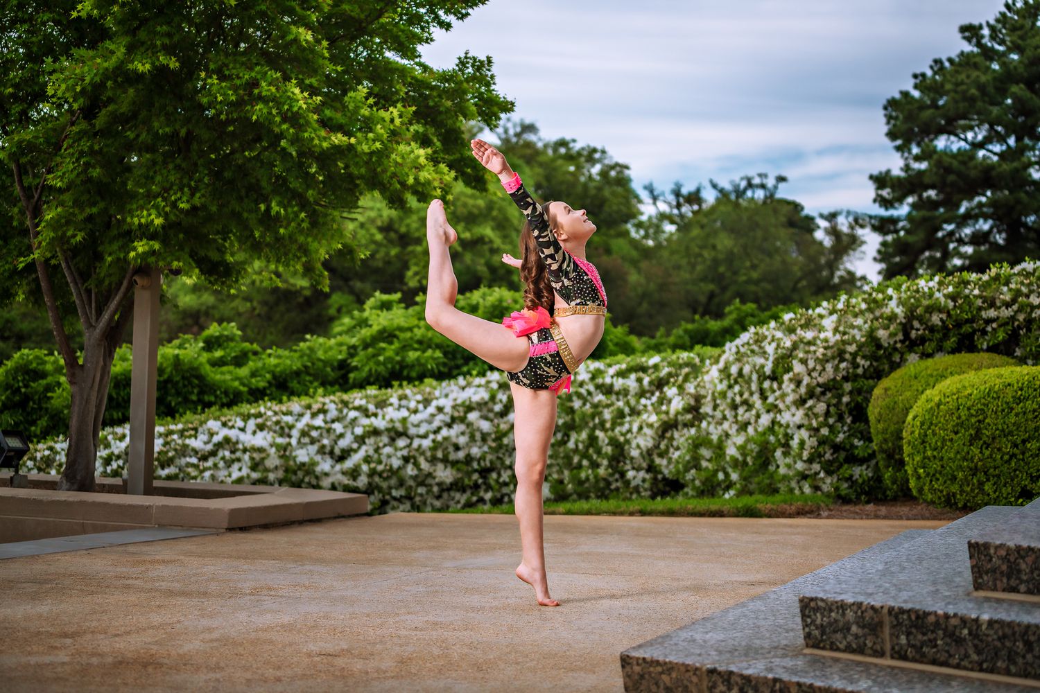 Young female dancer performing a high leg hold in a pink costume outdoors in a garden setting.