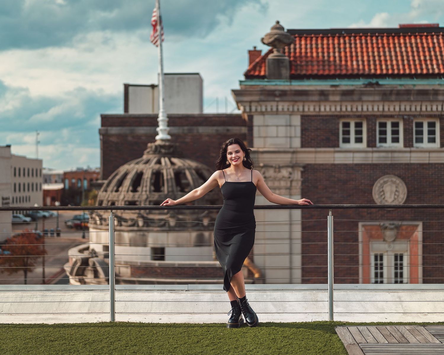 A woman in a black dress poses confidently on a rooftop with a historic building backdrop.
