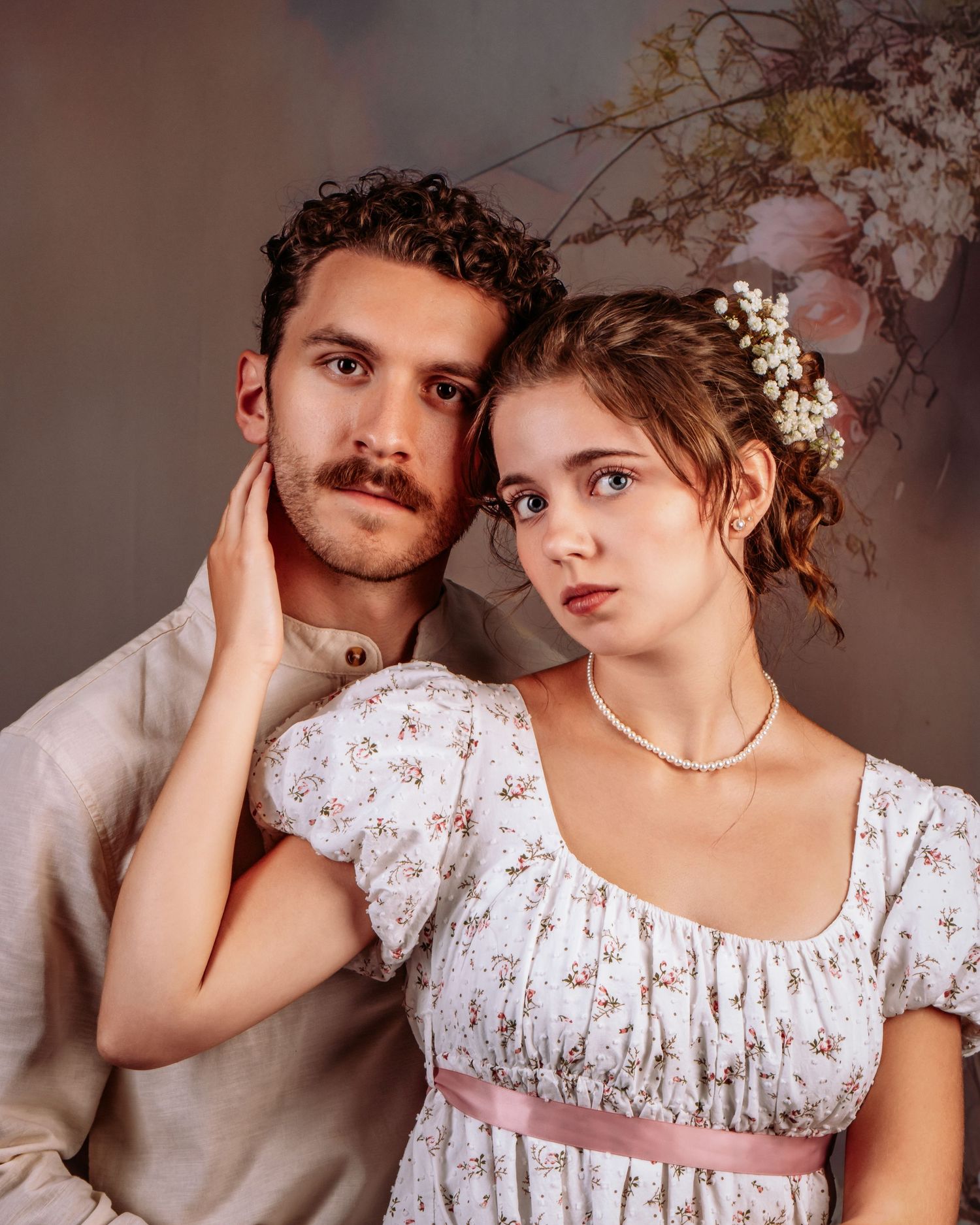 A couple in Regency-era costumes pose together with floral decorations, the woman in a pink-sashed dress.