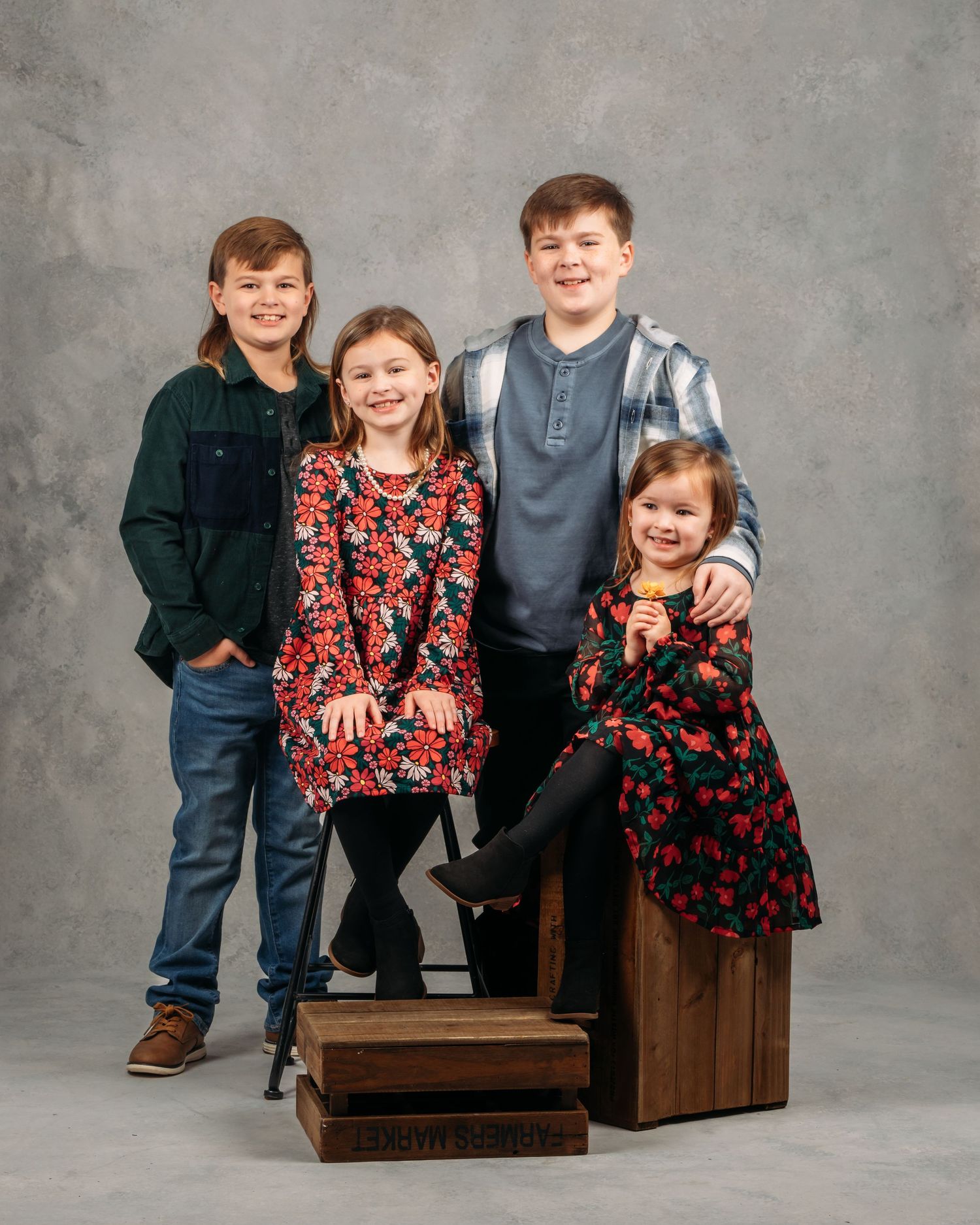 Four children pose together for a studio portrait on wooden crates with gray backdrop.