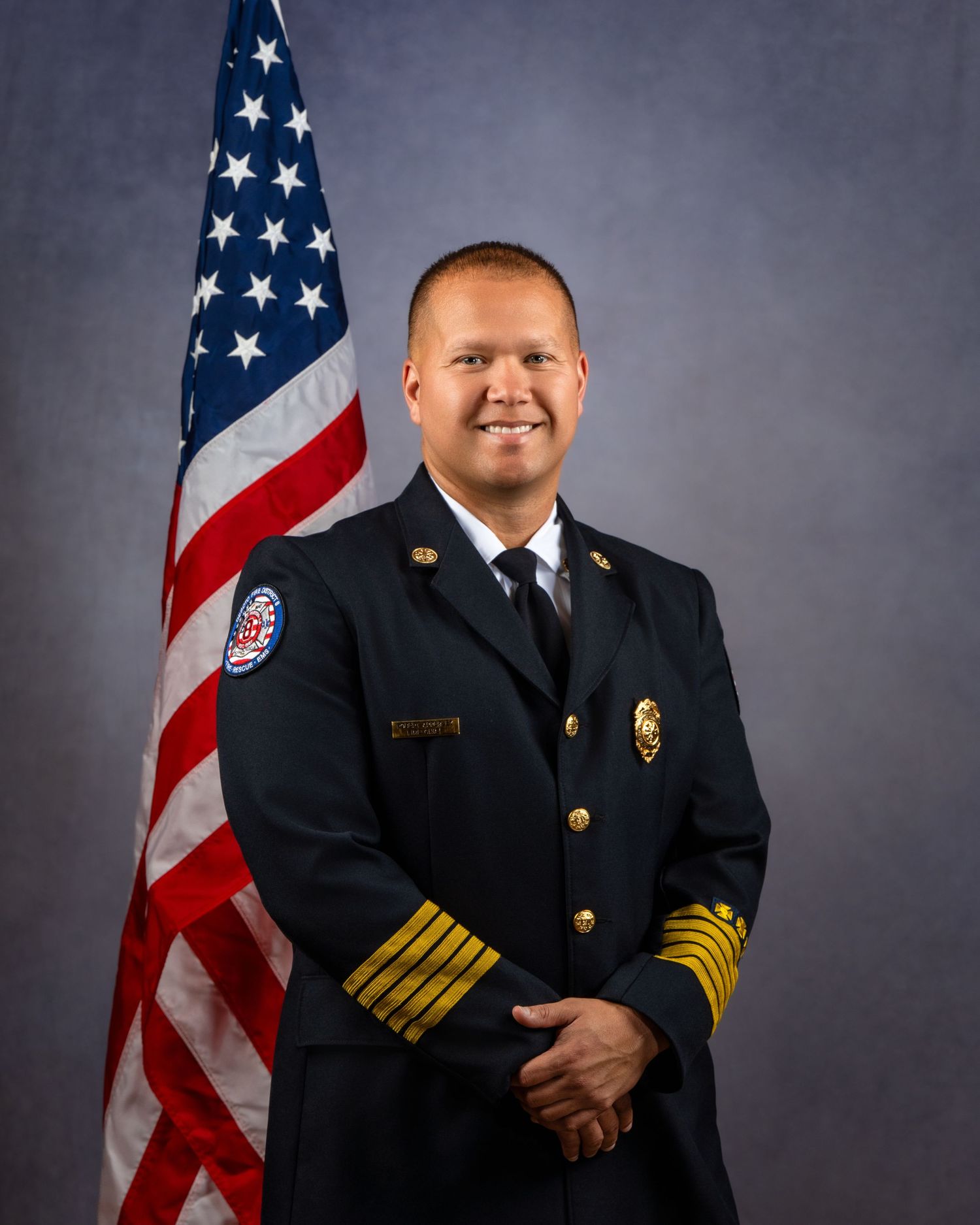 Fire department chief in dress uniform with gold insignia stands before American flag in official portrait photo.