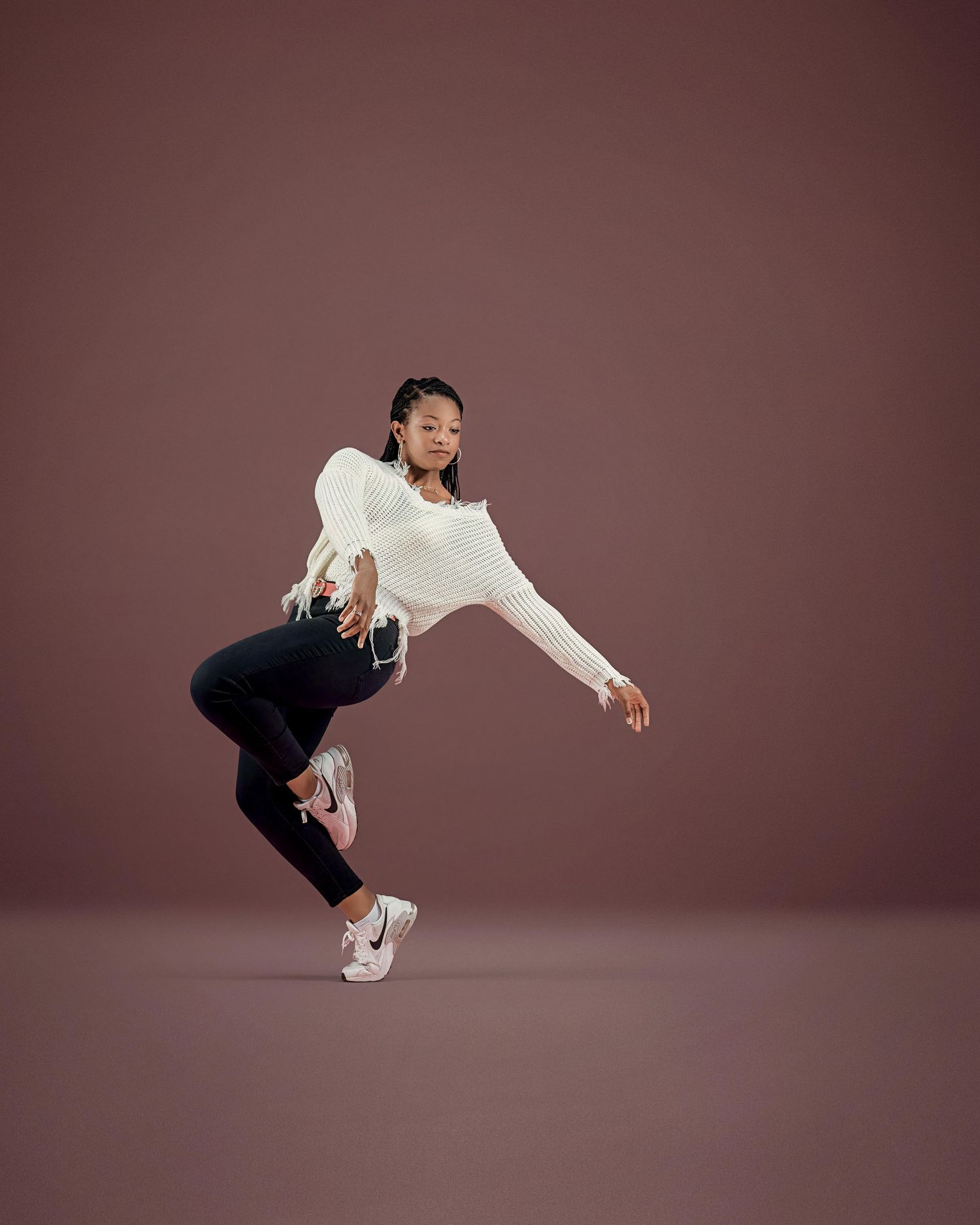 A young Black woman dances dynamically in a white knit sweater, black leggings, and Nike sneakers against a mauve background.