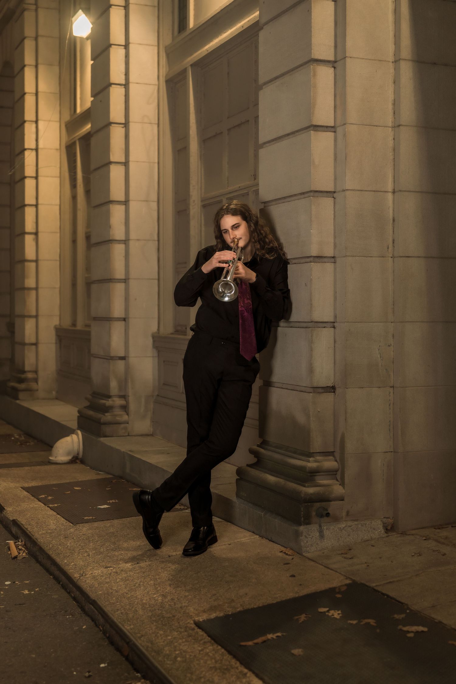 A musician in black attire plays trumpet outdoors at night, leaning against a stone building wall.