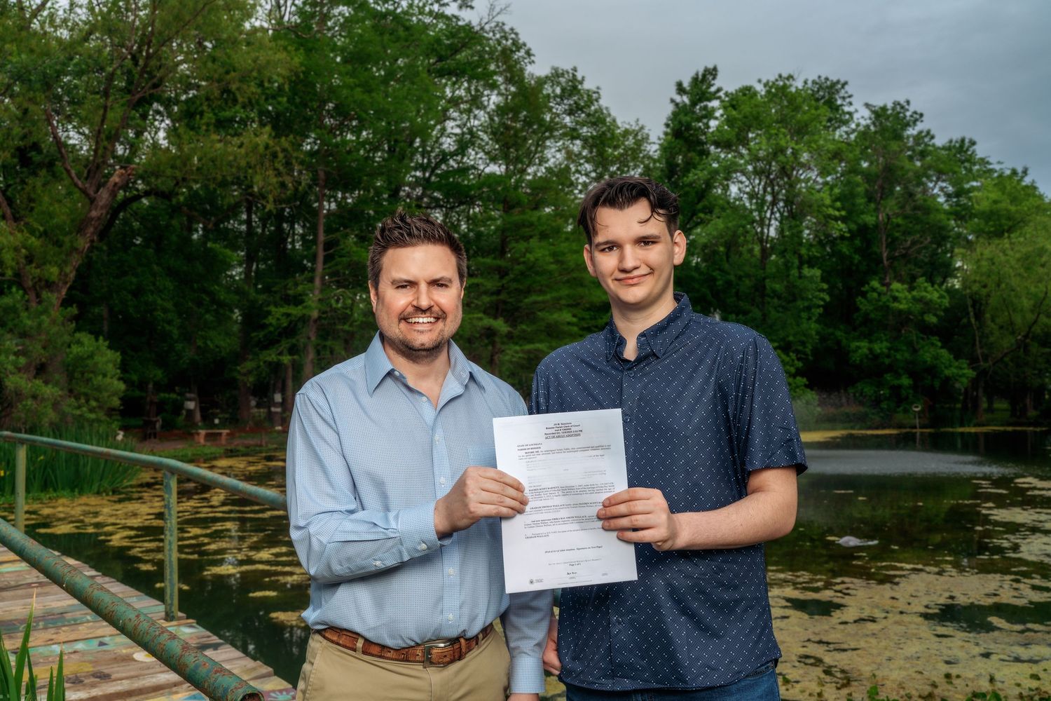 Two men smile proudly while holding a certificate near a scenic pond surrounded by trees.