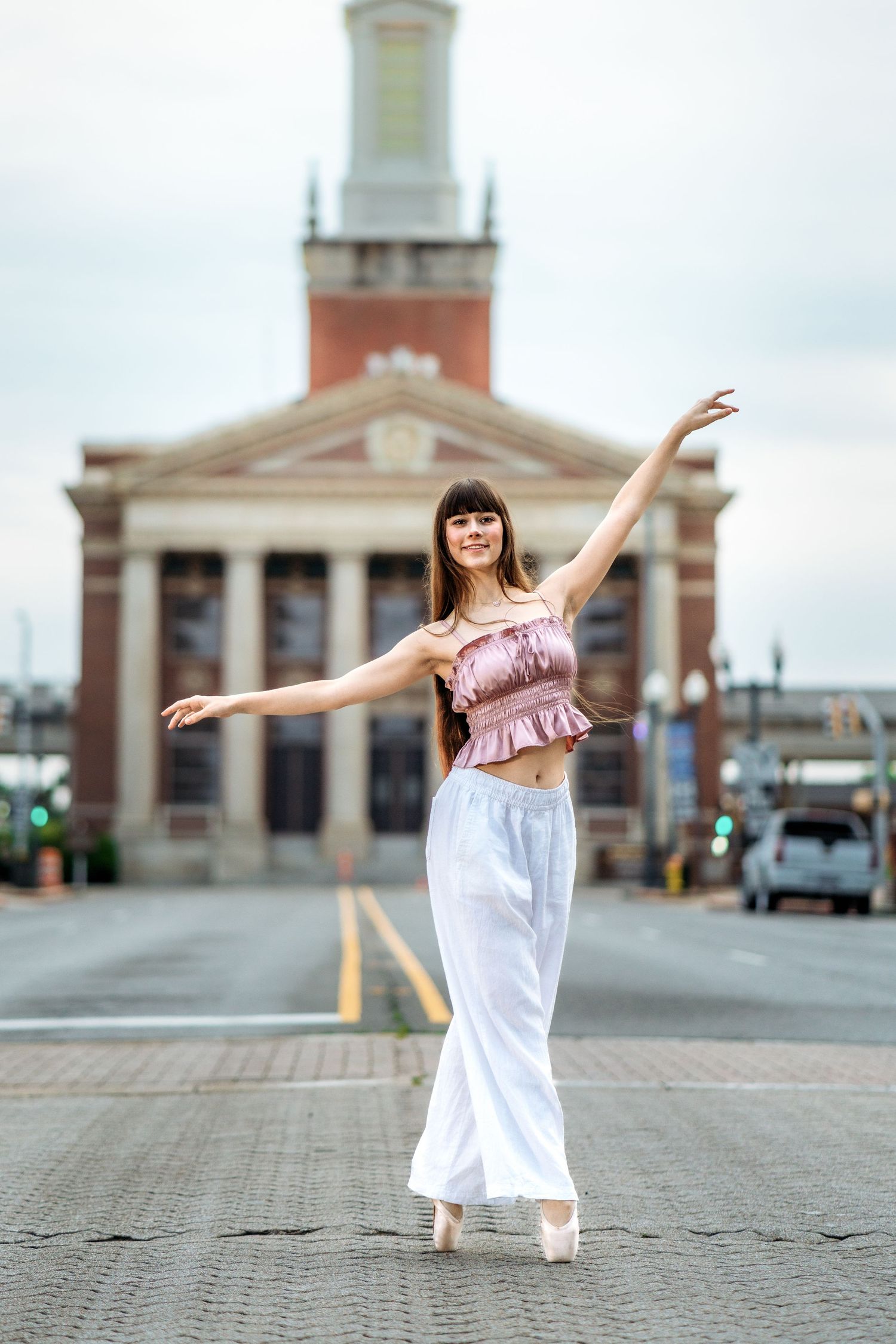 A ballerina poses gracefully in white pants and pink top on a city street, with a historic brick building behind her.