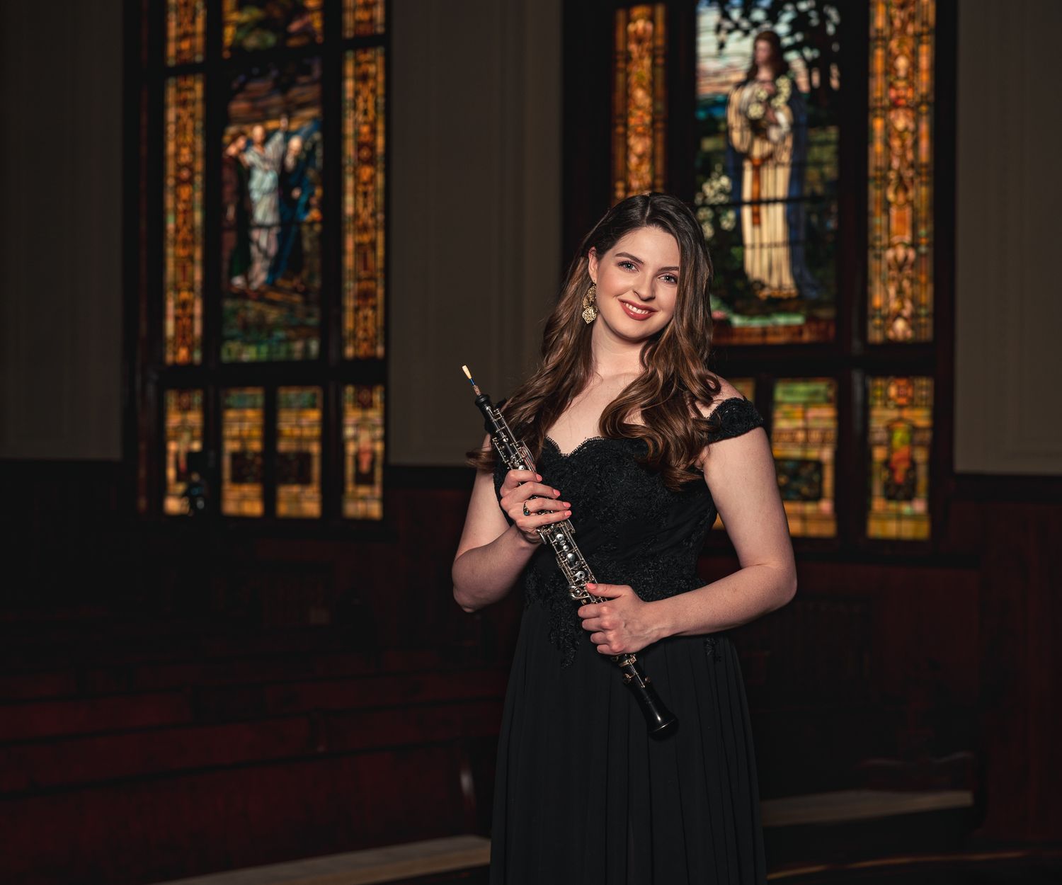 Smiling woman in black gown holding clarinet before stained glass windows.