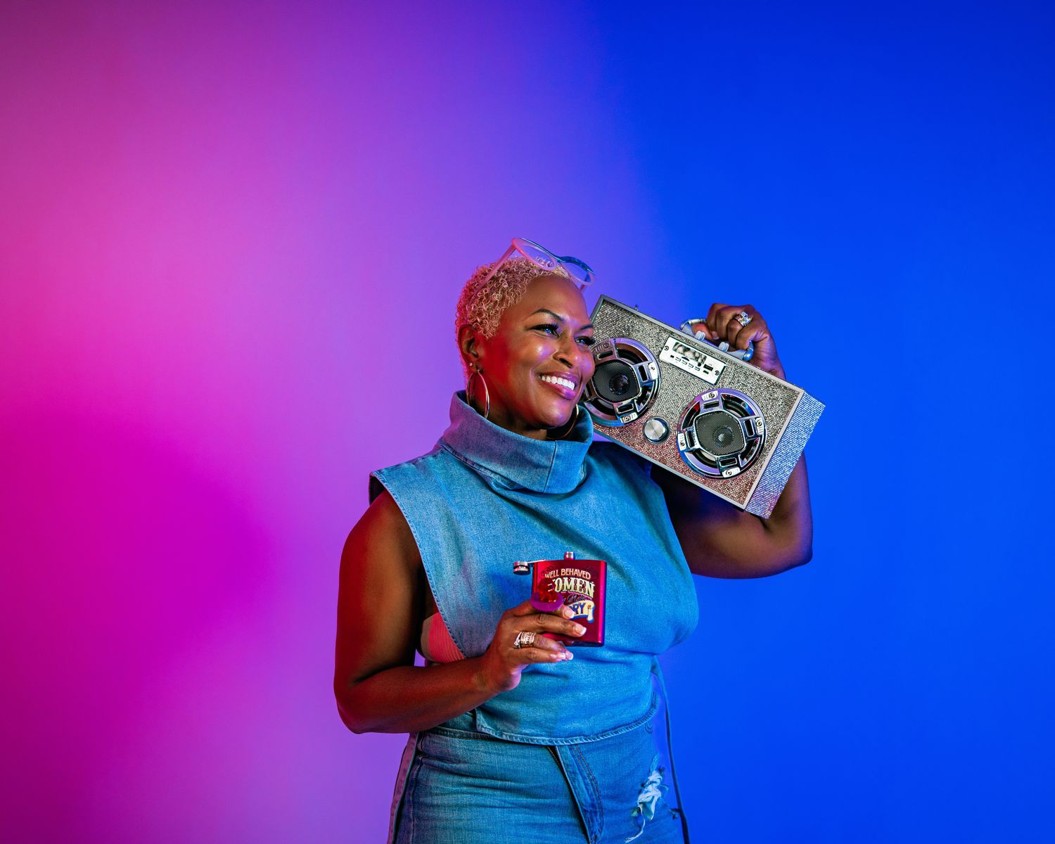 Woman with short pink hair holding a boombox on her shoulder in colorful neon pink and blue studio lighting.