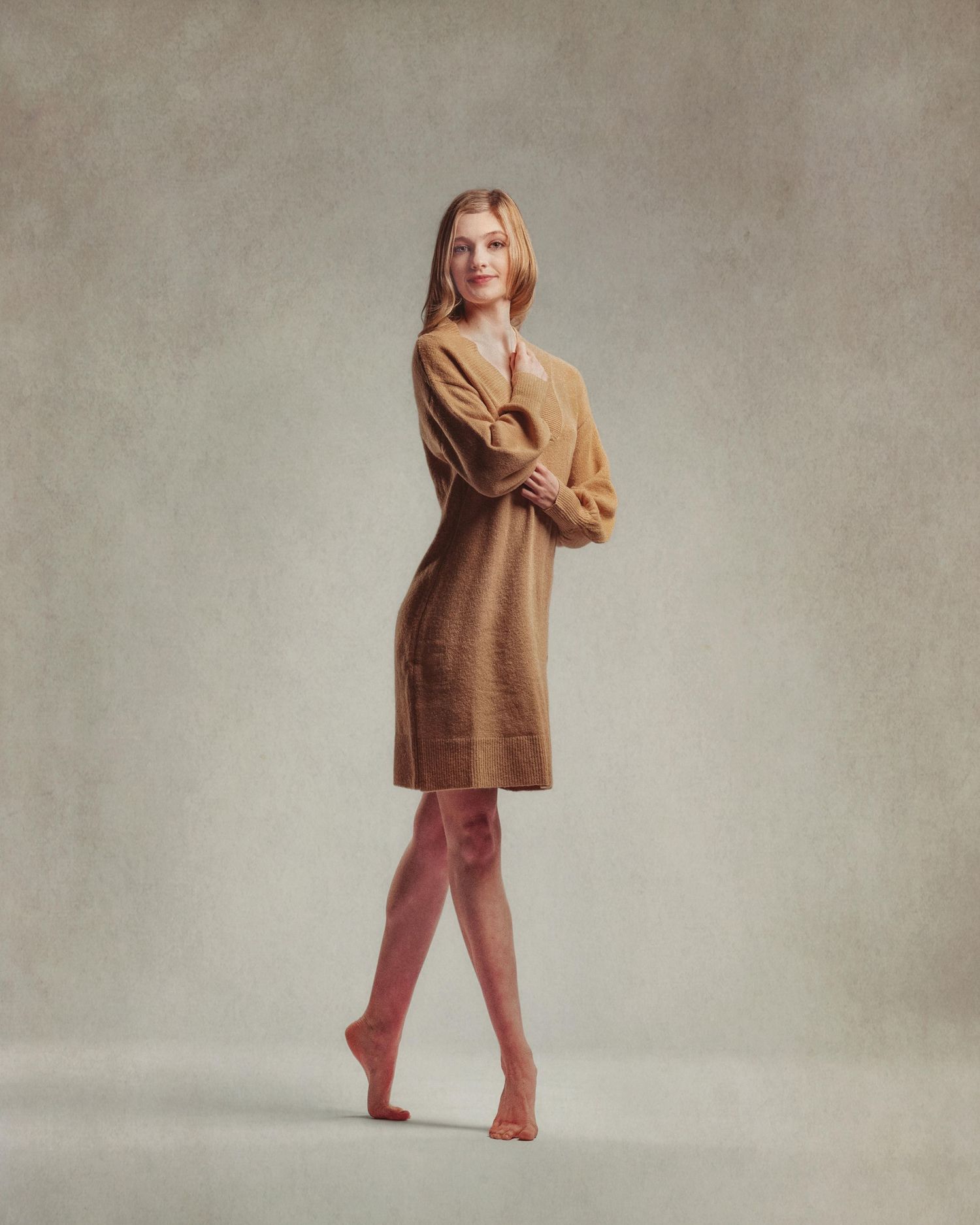 Woman in camel knit sweater dress and nude heels posing gracefully against a neutral gray background.