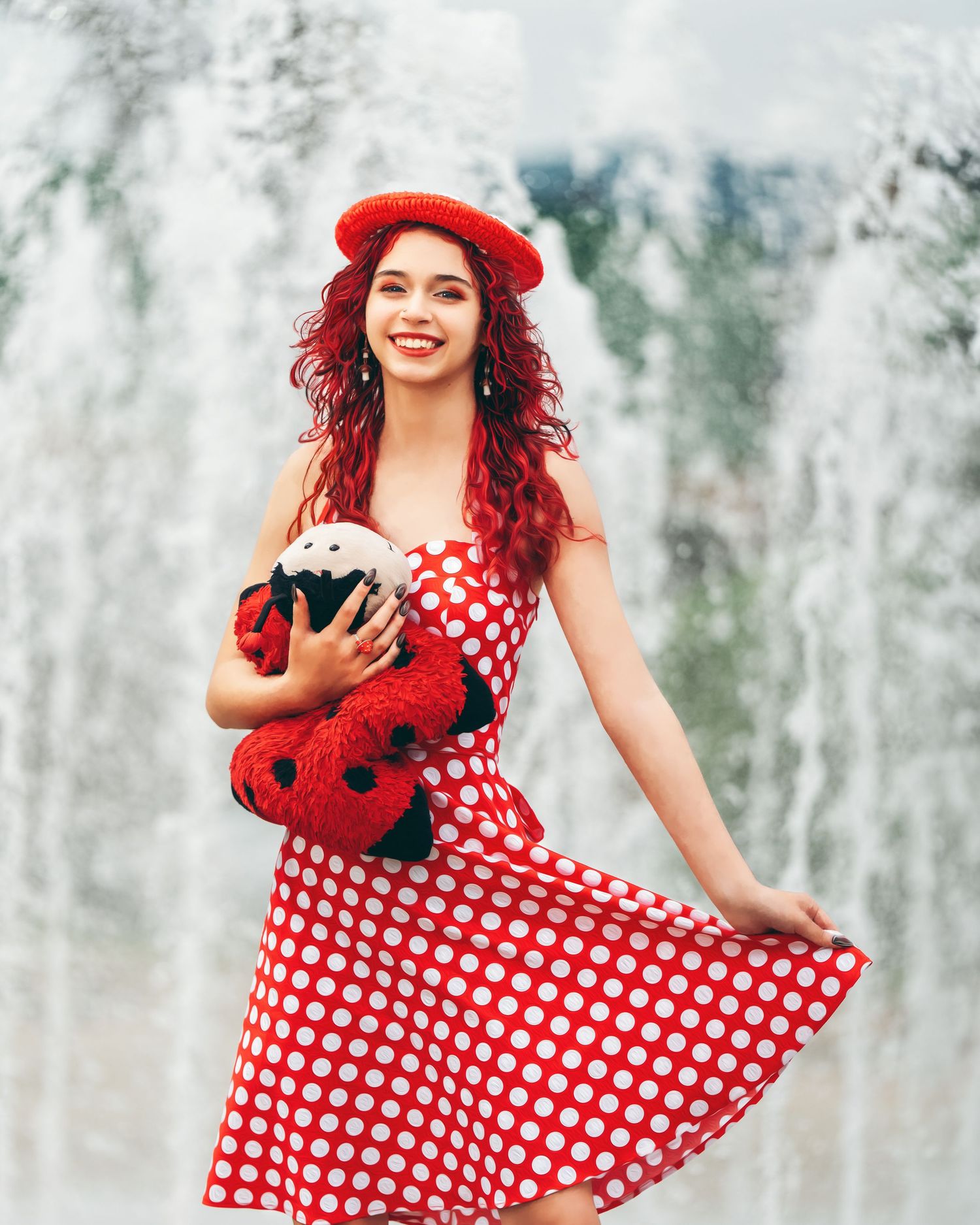 Young woman in red polka dot dress and beret holds ladybug plush toy, smiling by fountain.