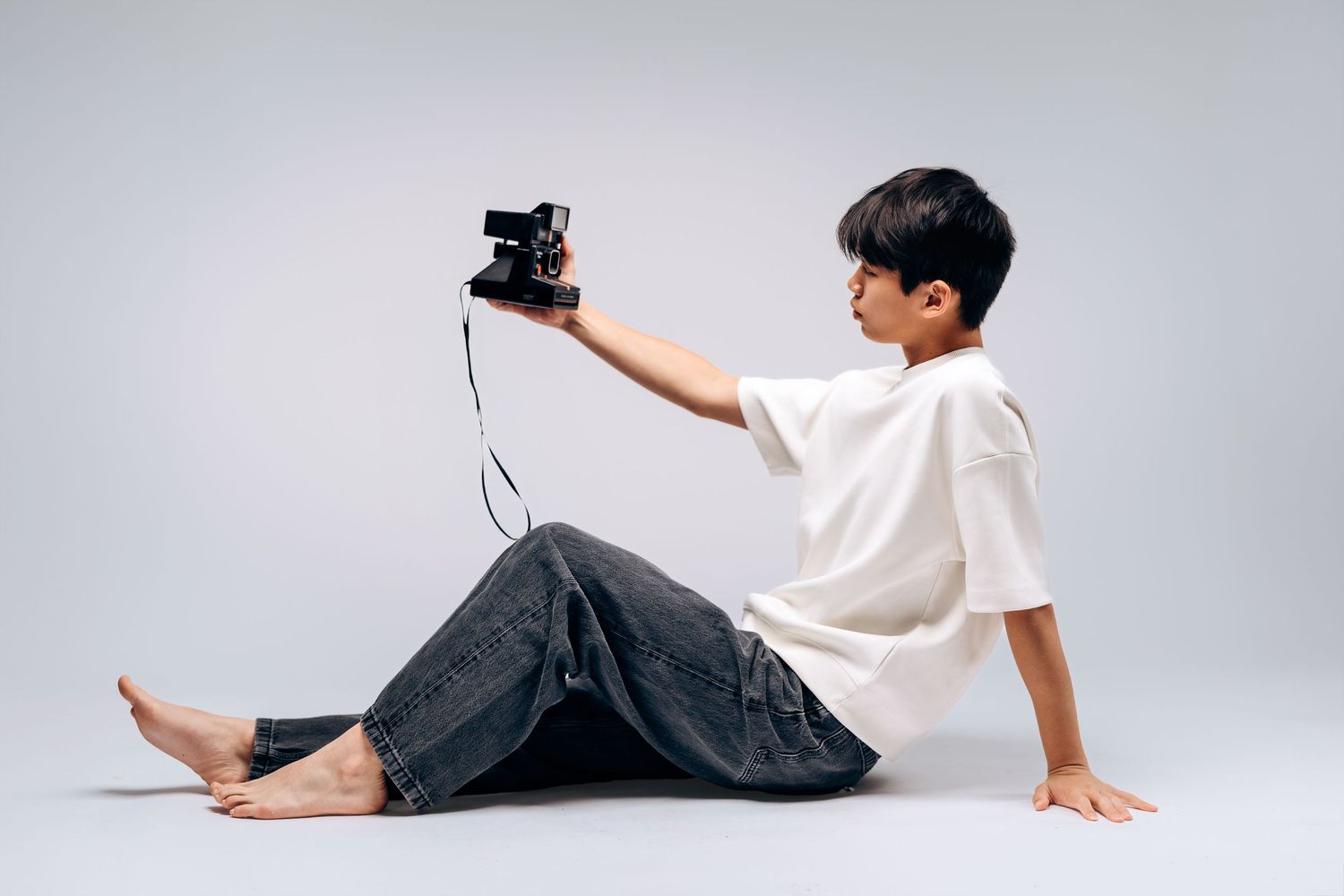 A barefoot boy in white shirt and jeans sits on floor, holding a vintage camera up for a selfie.