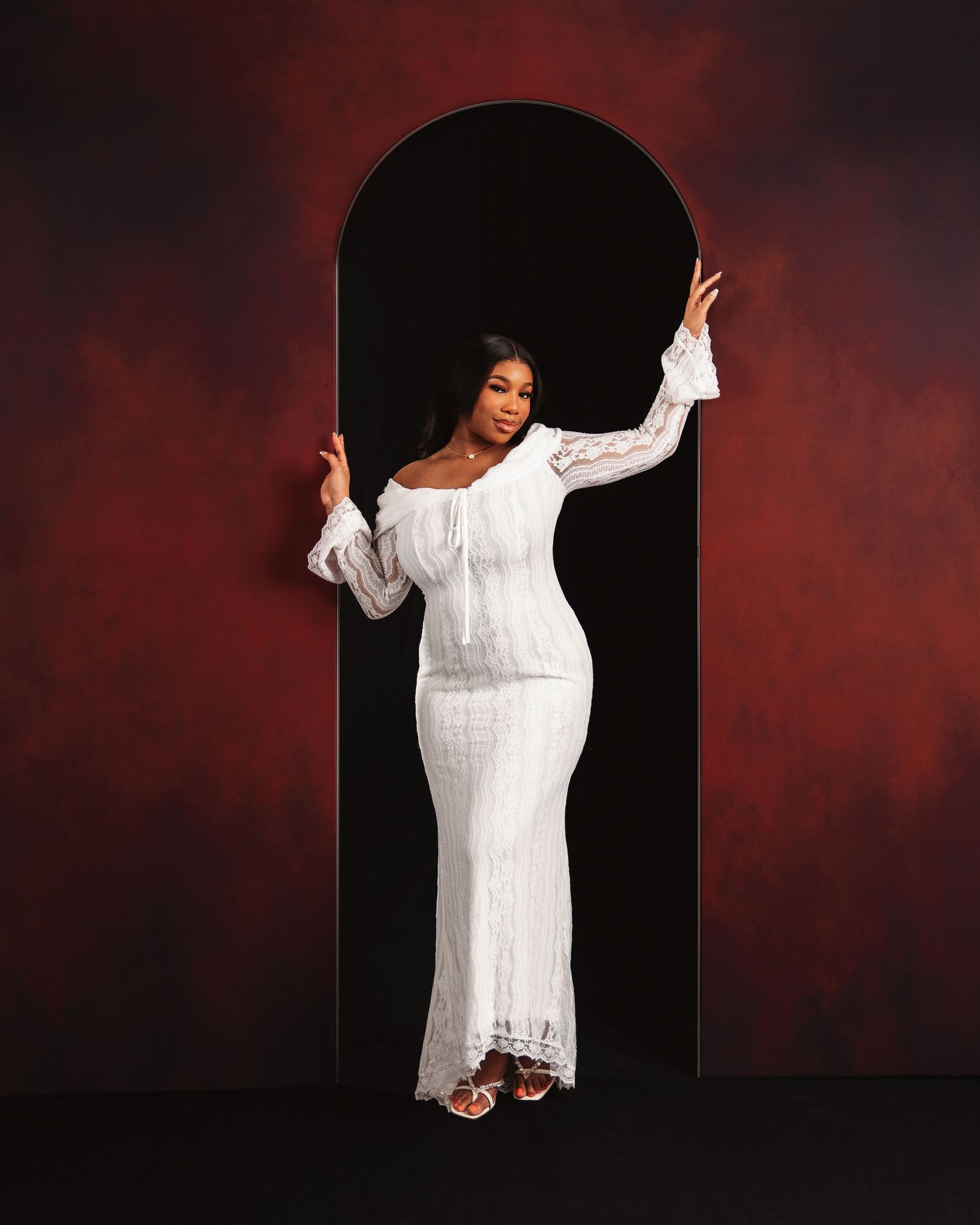 Woman in white lace maxi dress posing in black arch doorway against dramatic dark red background.
