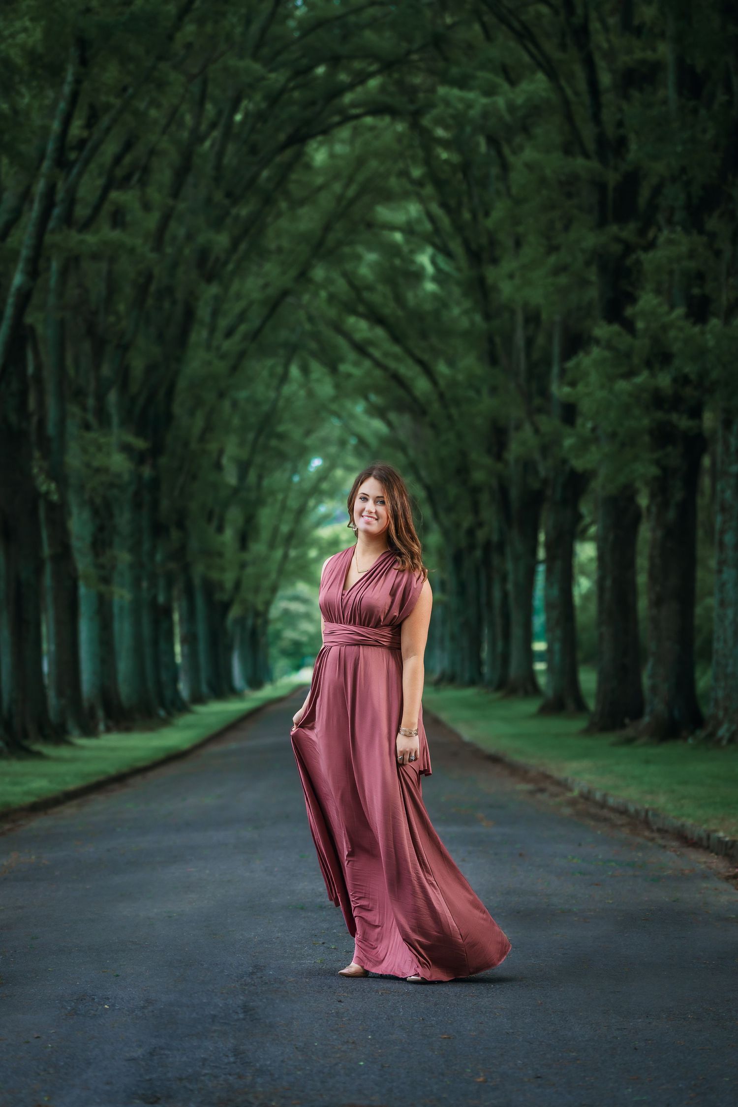 Woman in flowing mauve gown stands on road beneath dramatic arching green tree tunnel.