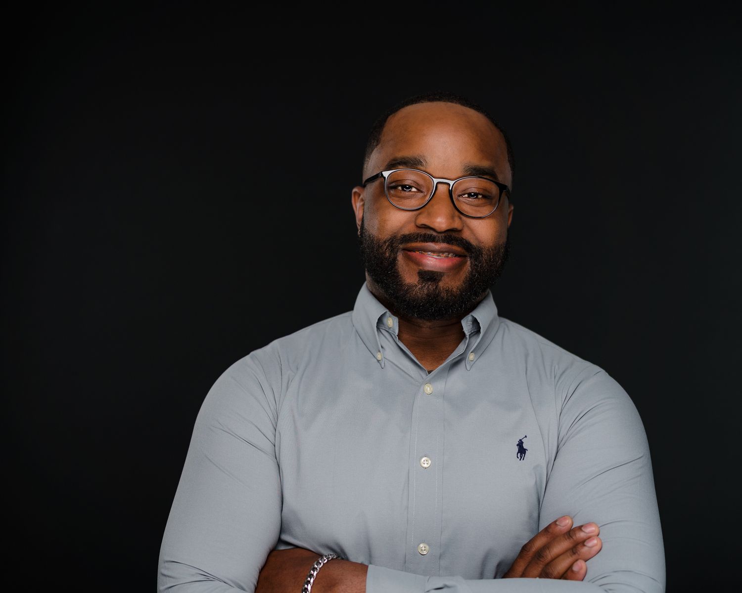 Smiling Black man in gray polo shirt with crossed arms posing for professional headshot.