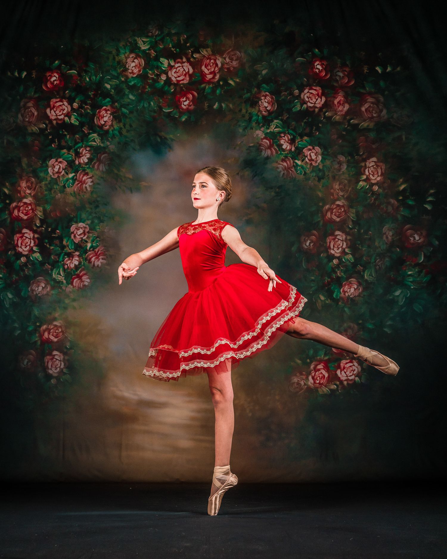 Young ballerina in red tutu dress performs arabesque on pointe shoes with floral backdrop.