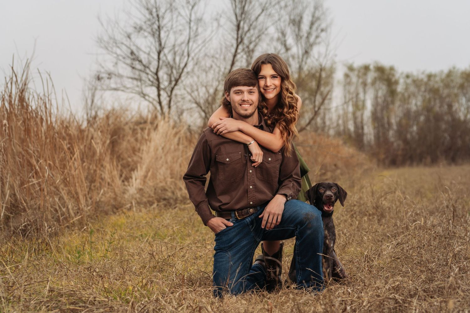Smiling couple posing with their black Labrador dog in a rustic autumn field.