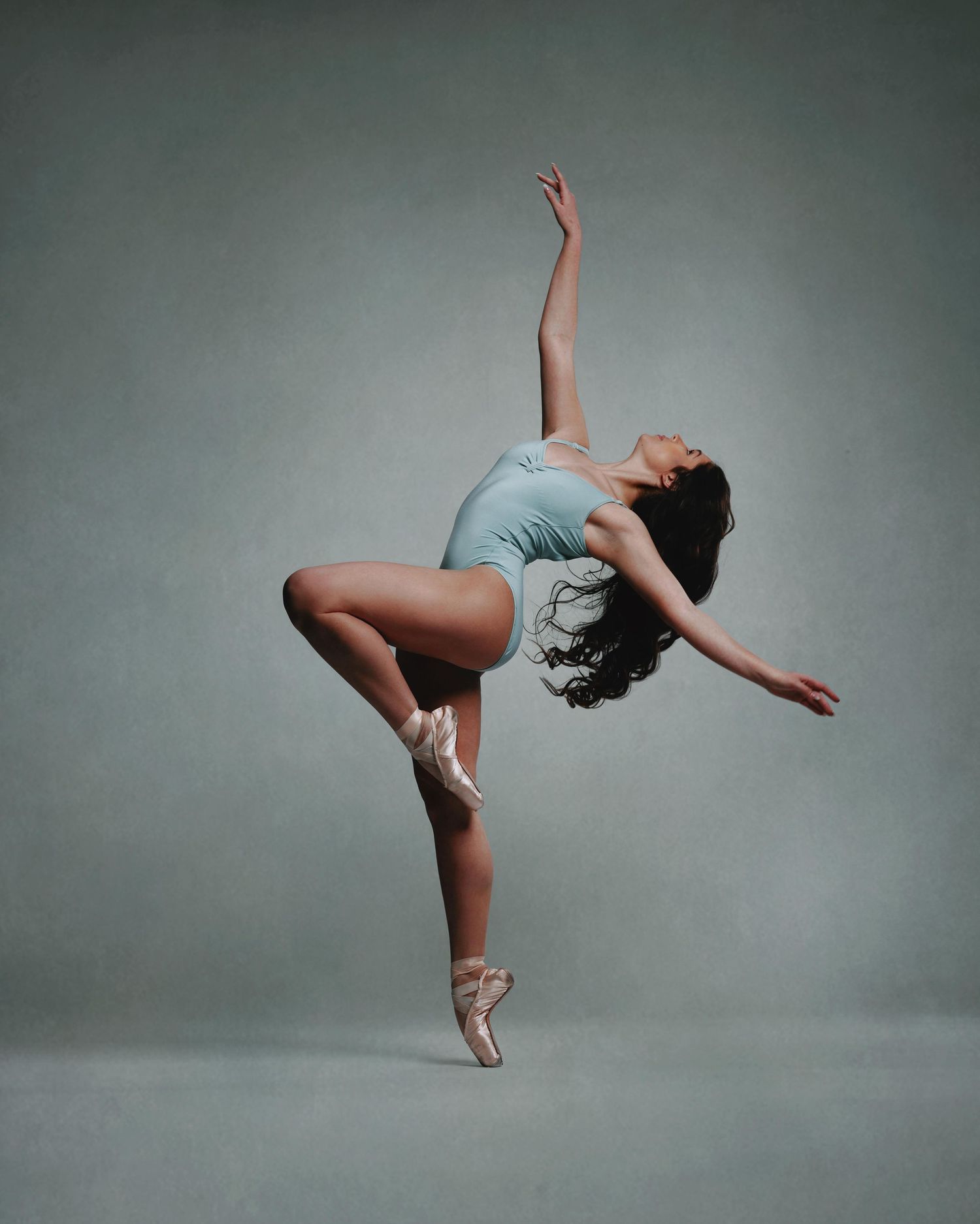 A ballet dancer in a light blue leotard and pointe shoes strikes an elegant arabesque pose against a gray studio backdrop.
