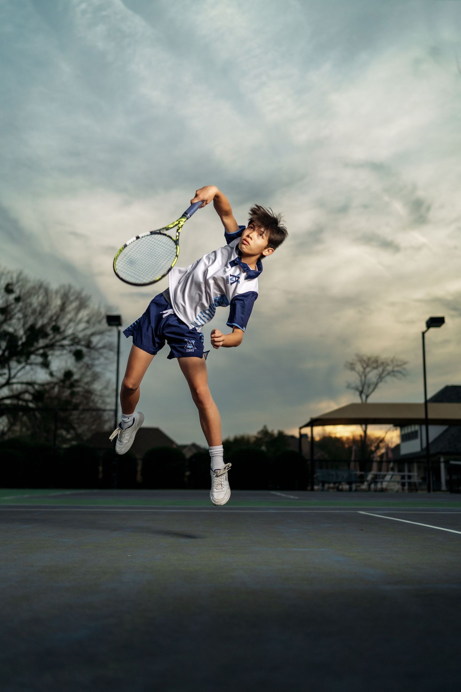 Young tennis player jumping mid-air while serving on an outdoor court at dusk.