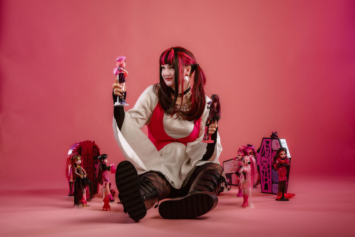 Young woman in costume sits on pink background holding Monster High dolls surrounded by collectible figures.