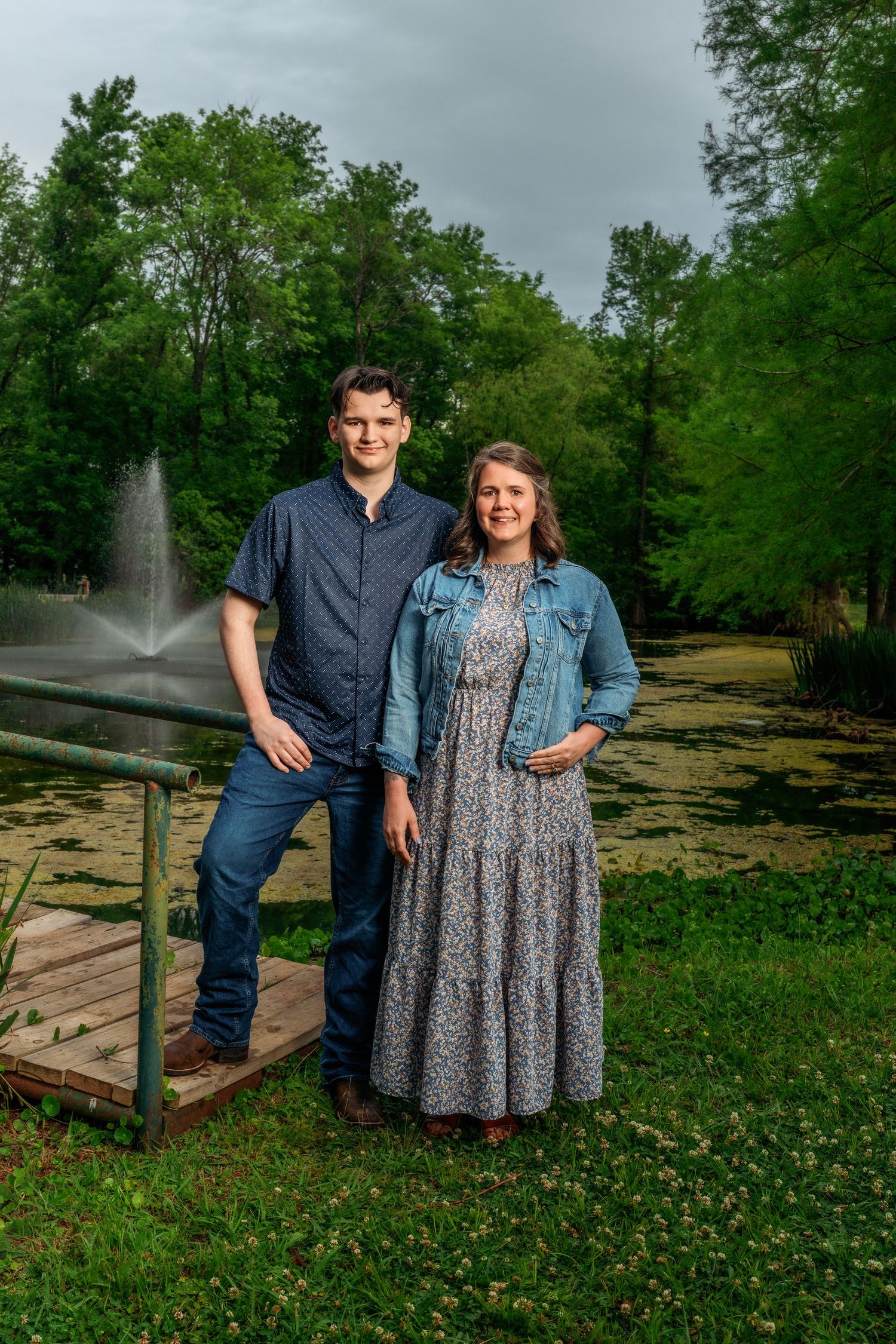 A couple stands together on a wooden dock beside a serene pond surrounded by lush green trees.