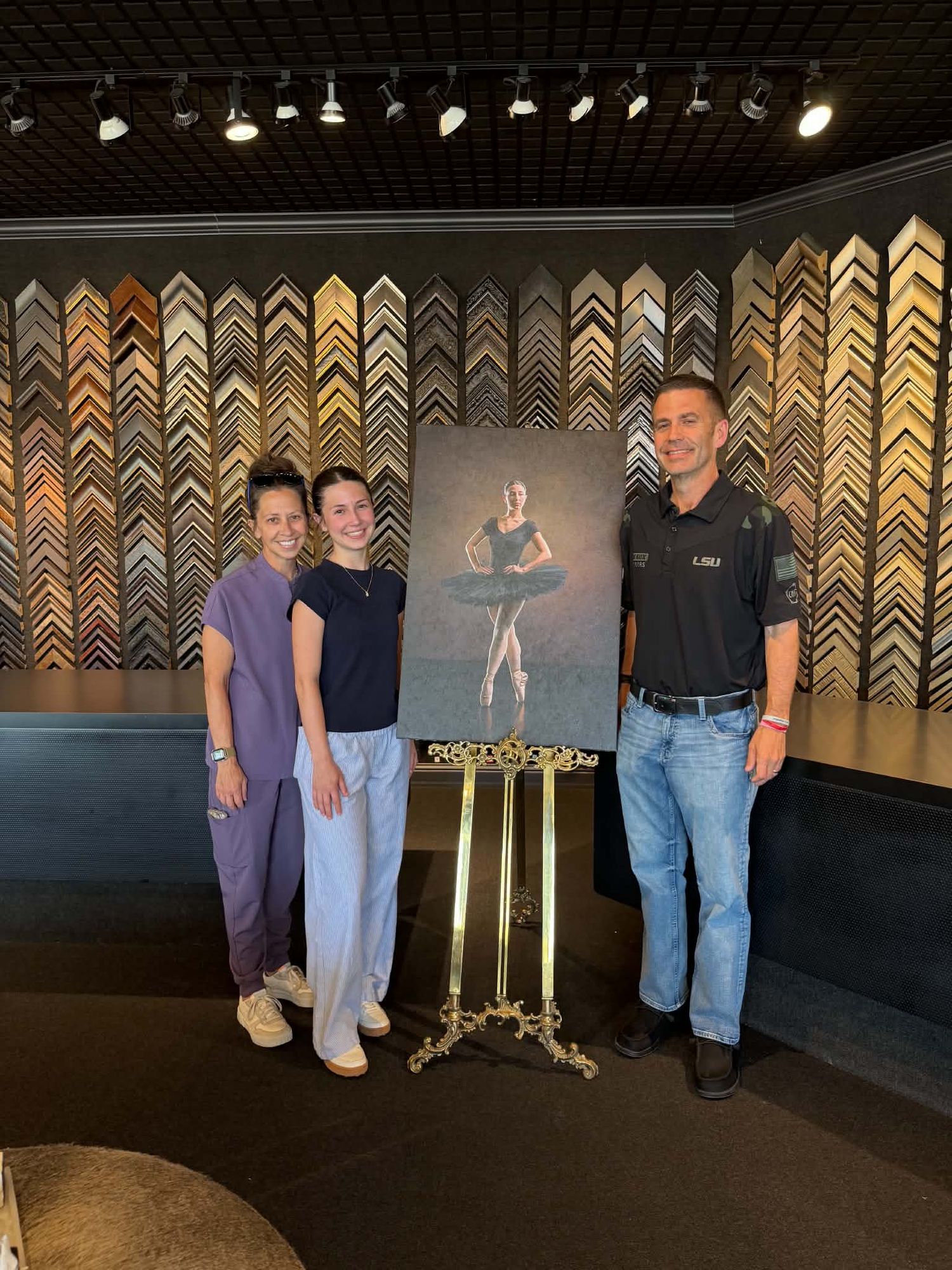 Three people pose beside a framed ballerina portrait on a gold easel in a frame shop.