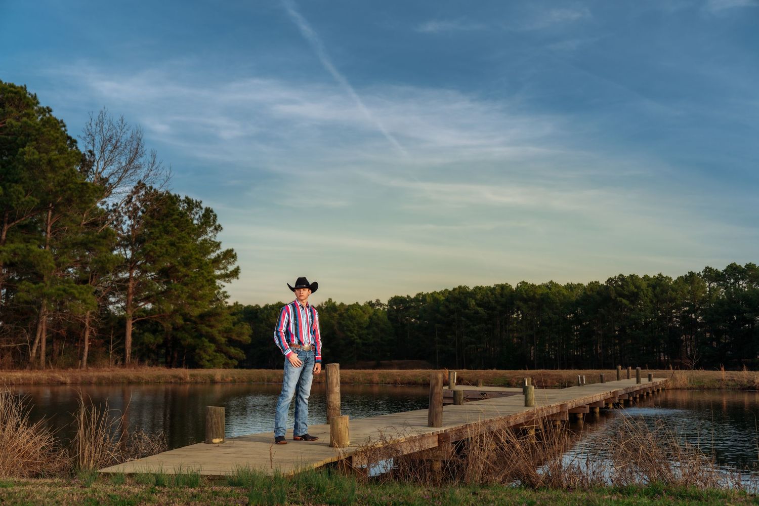 Cowboy in plaid shirt and hat standing on a wooden dock by a lake at sunset.