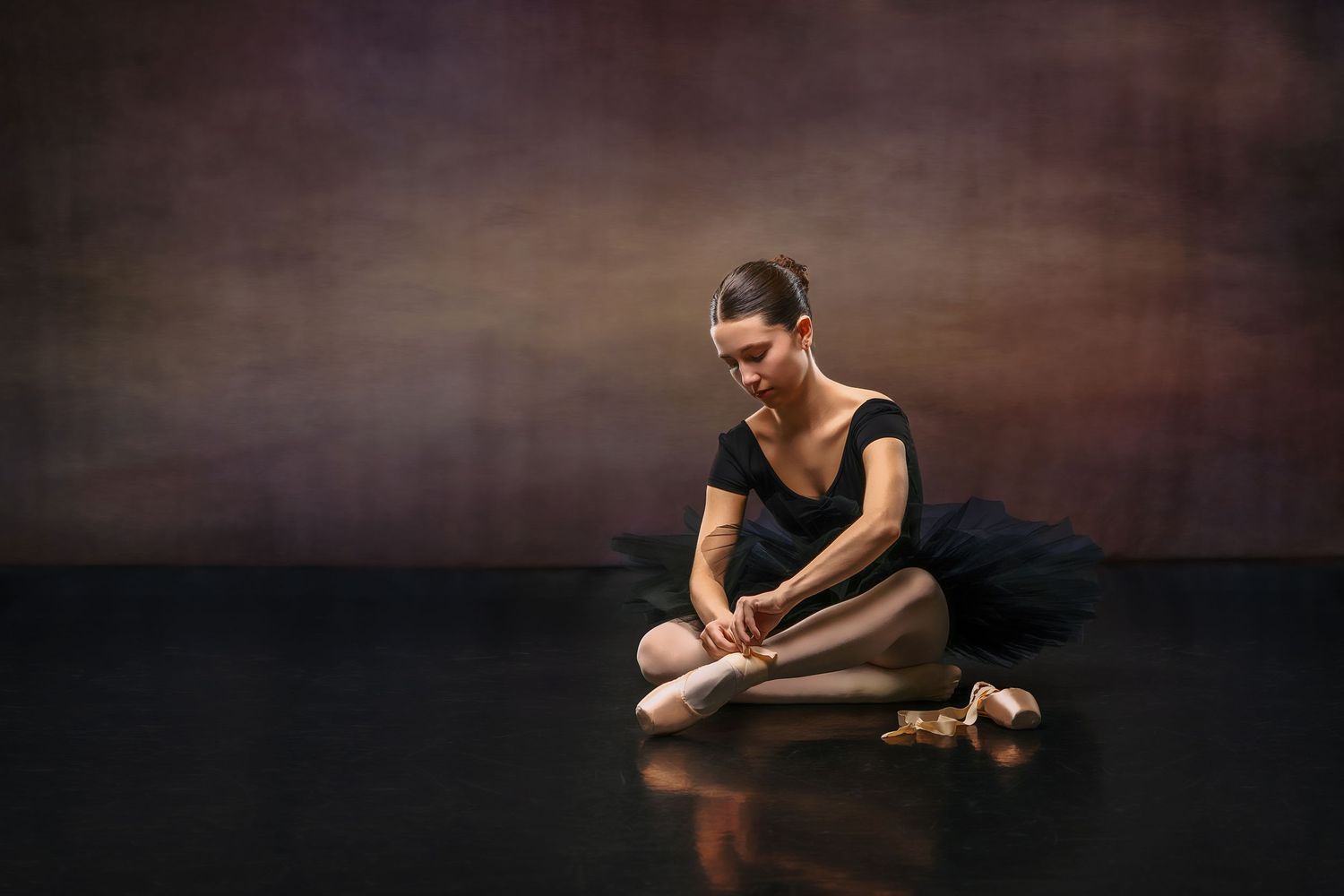 Ballerina in black tutu sitting on floor adjusting pointe shoes in a studio.