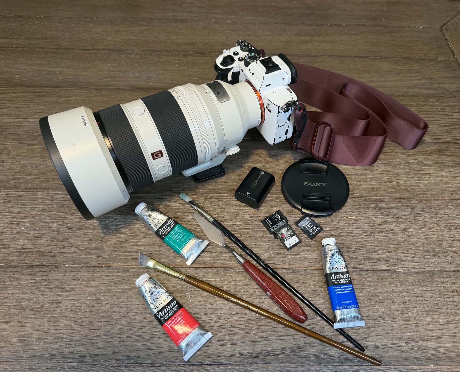 Sony mirrorless camera with telephoto lens, paint tubes, and brushes on wood surface.