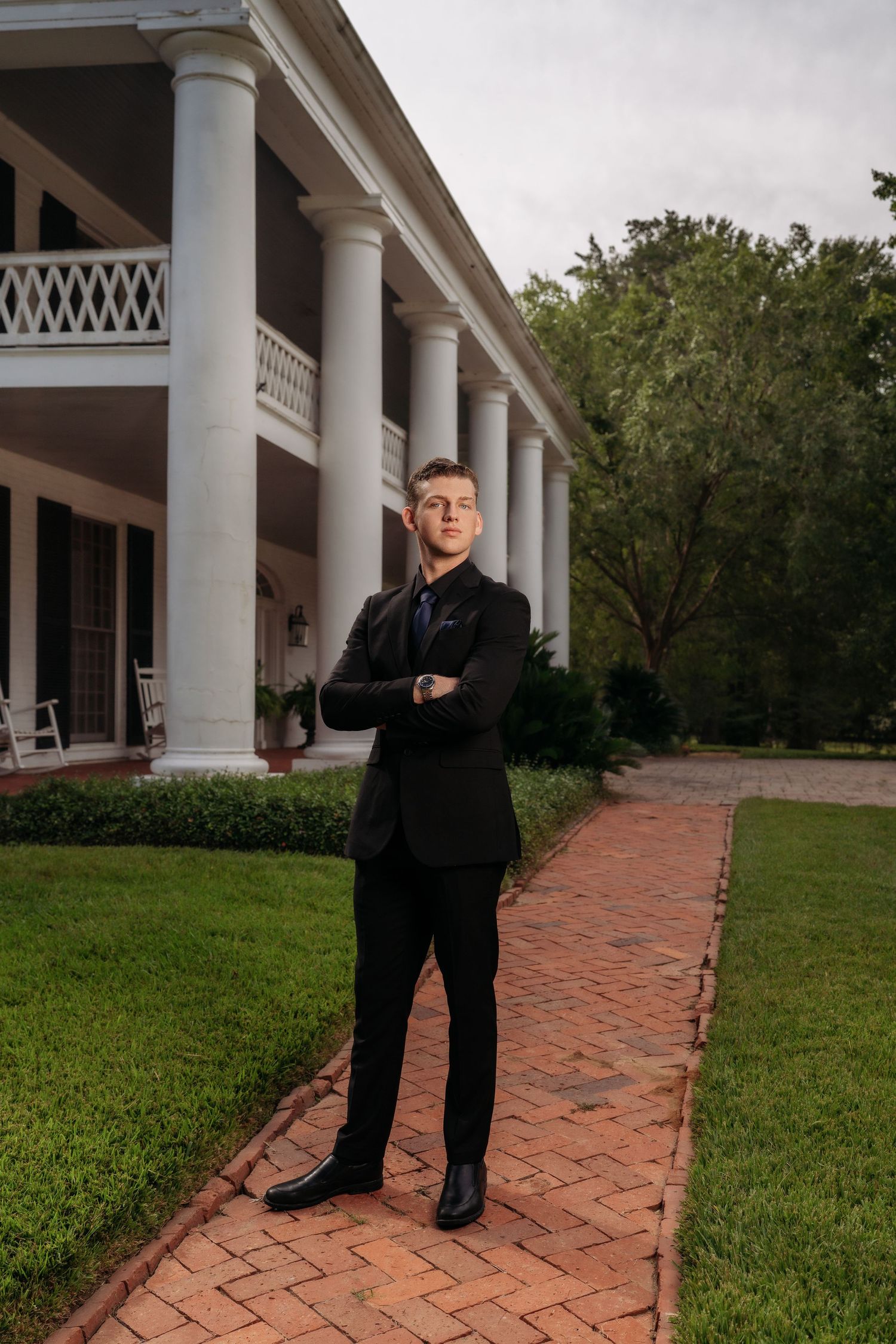 Young man in black suit standing arms crossed on brick path before grand white-columned Southern mansion.