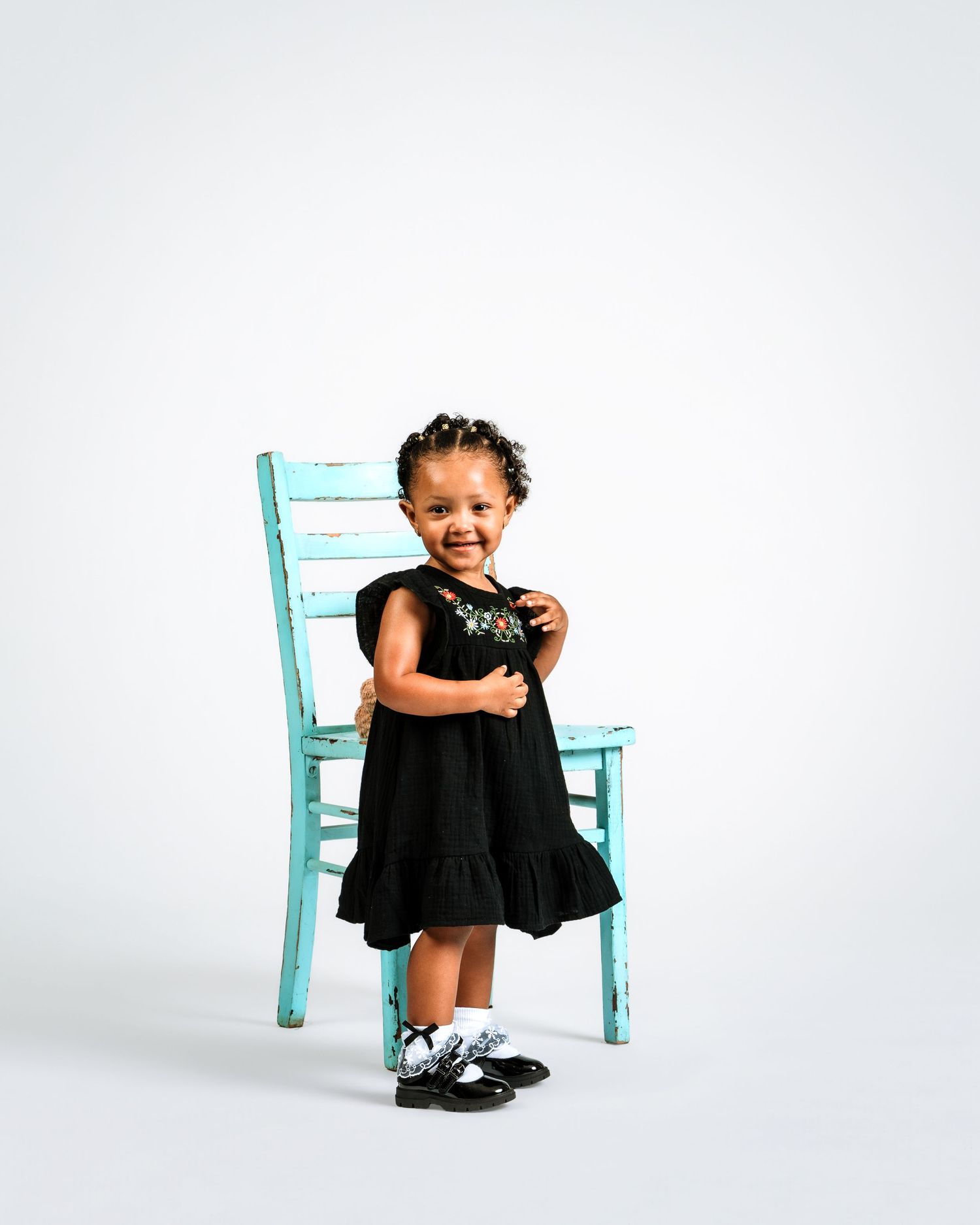 Adorable toddler girl in black dress smiles joyfully while leaning on a rustic turquoise chair.