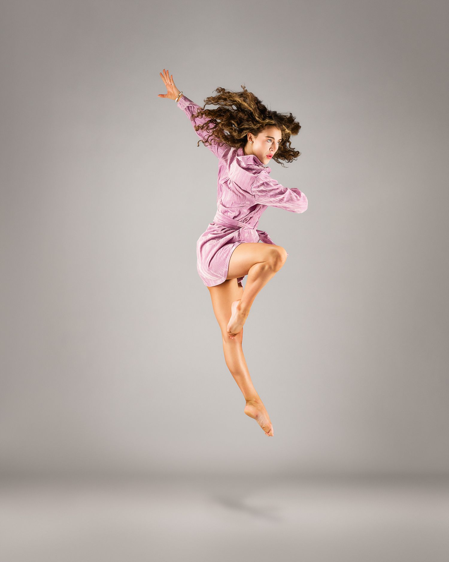 Female dancer mid-leap in a pink satin dress with flowing hair against a gray studio background.