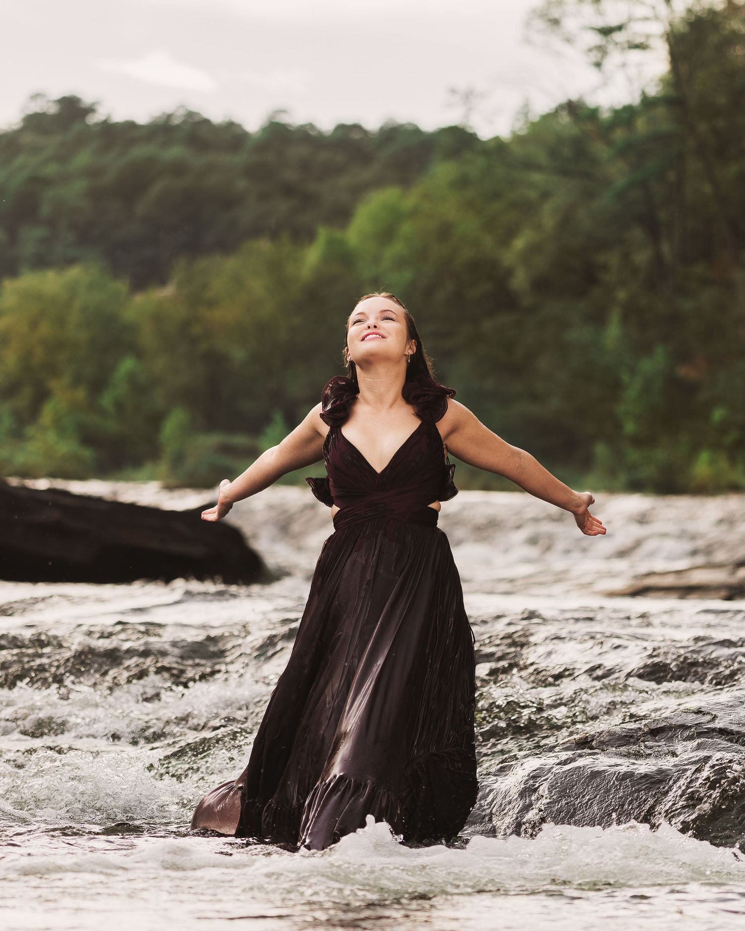 Woman in black dress standing in rushing river with arms outstretched among green trees.