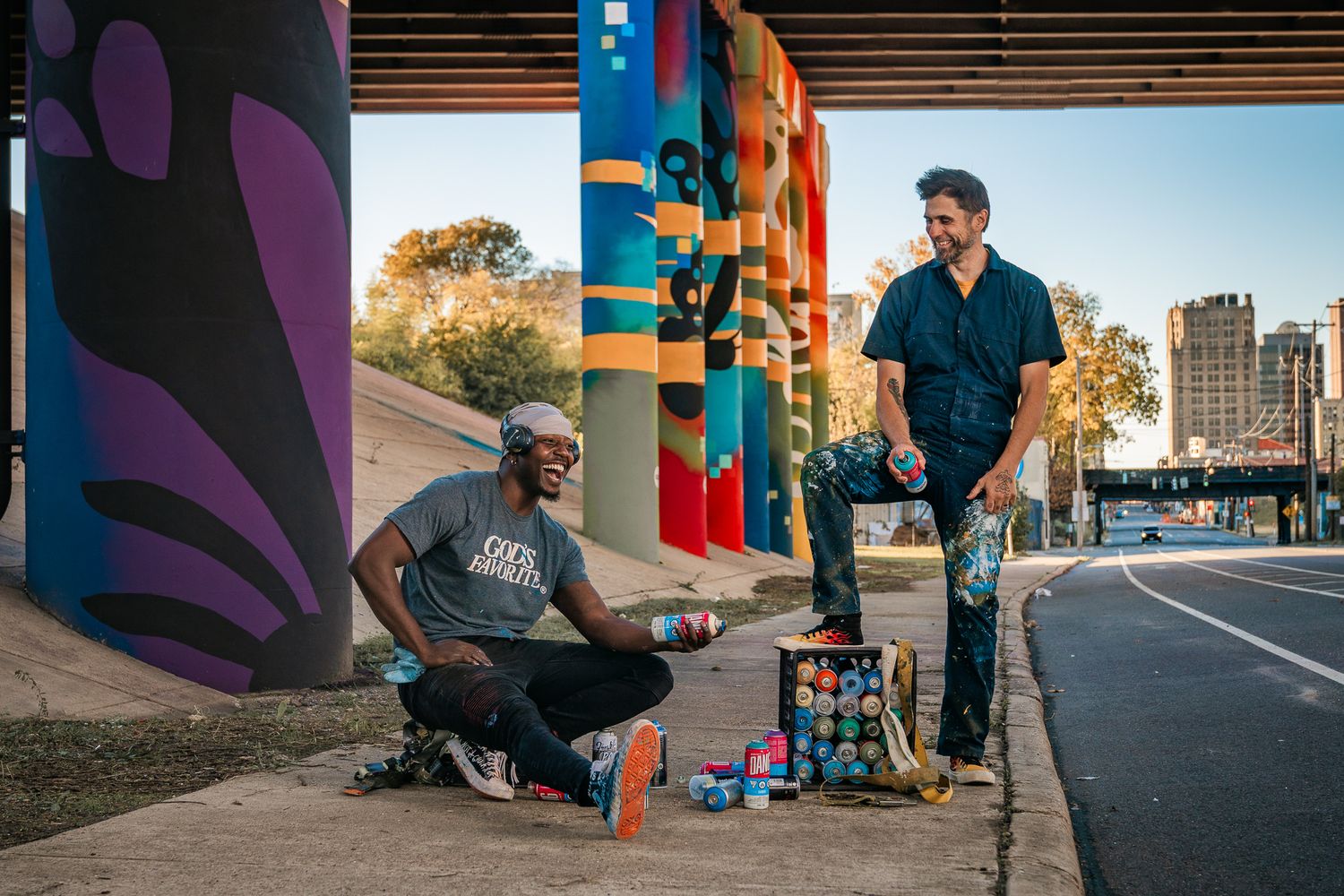 Two male street artists with spray paint cans working on colorful murals under an urban overpass.