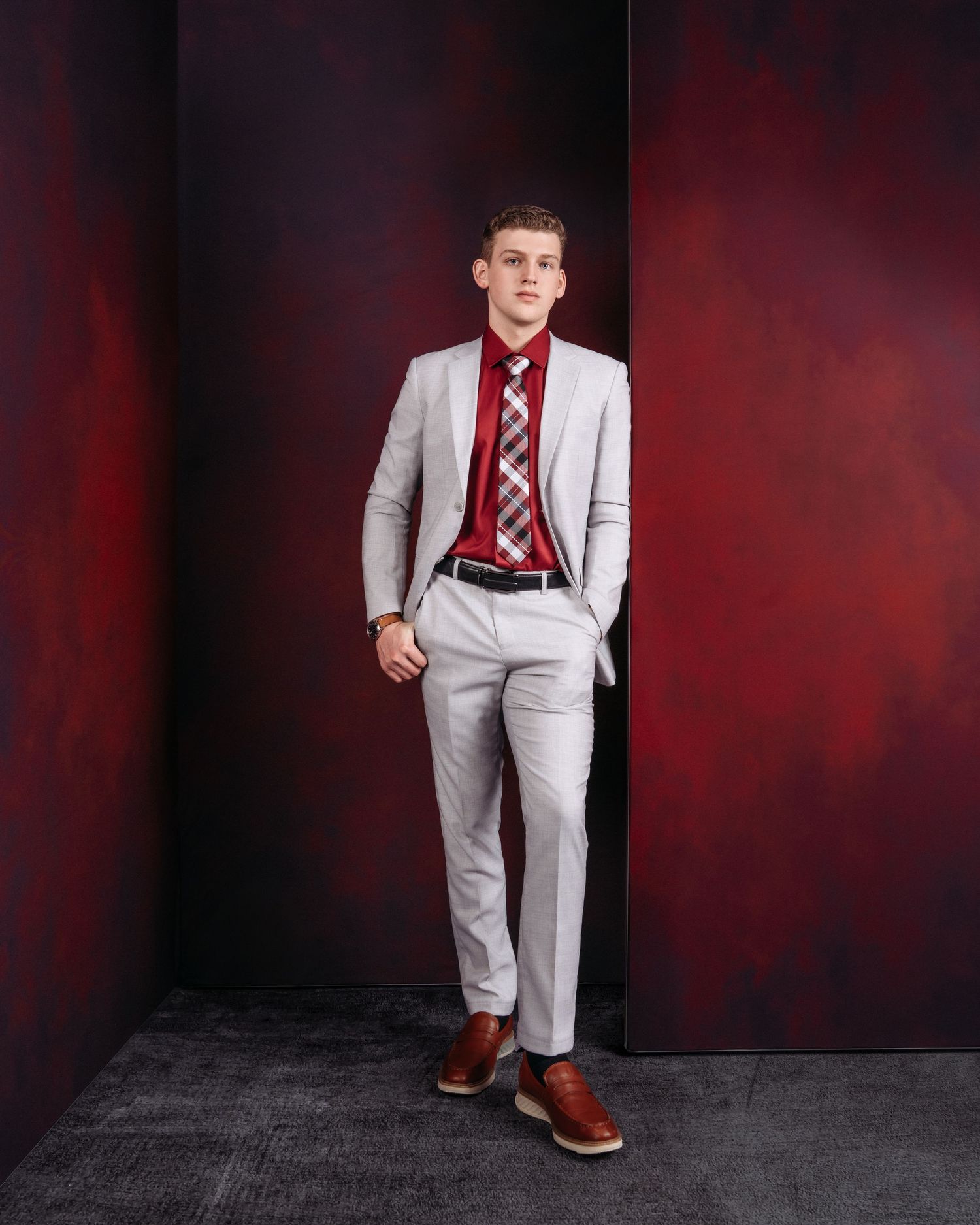Young man in light gray suit, red shirt, plaid tie, and brown dress shoes poses against a dark red backdrop.