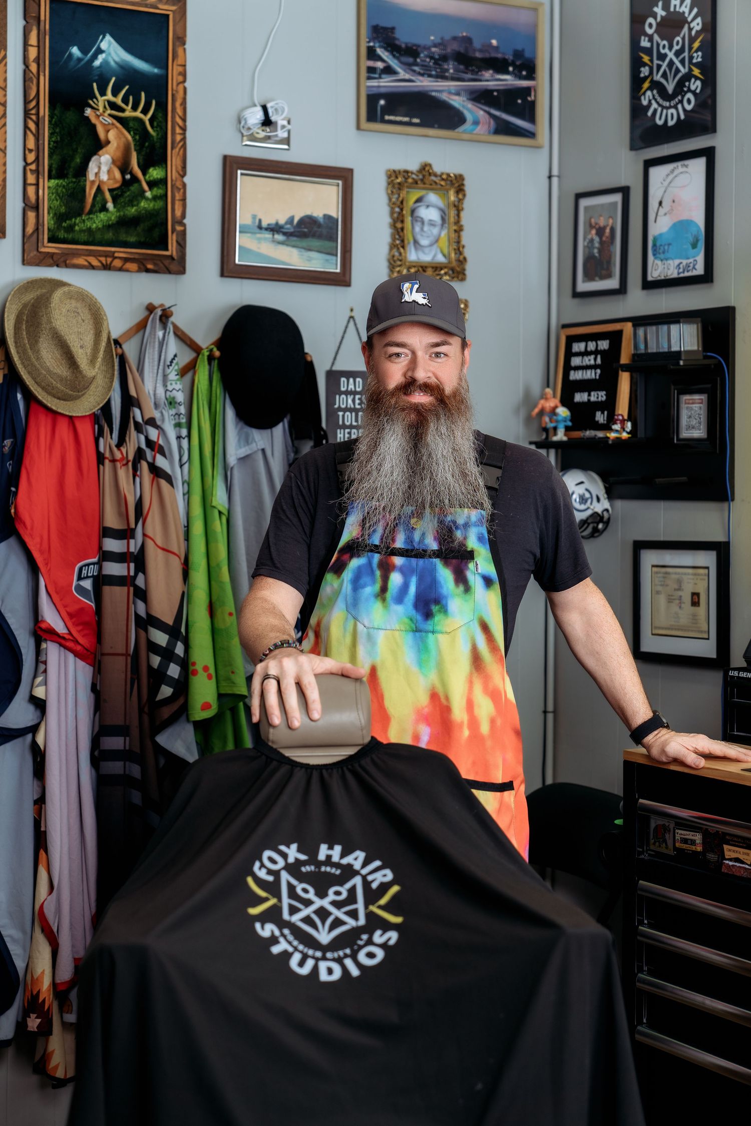 Bearded barber in tie-dye apron poses at Fox Hair Studios chair surrounded by eclectic wall art.