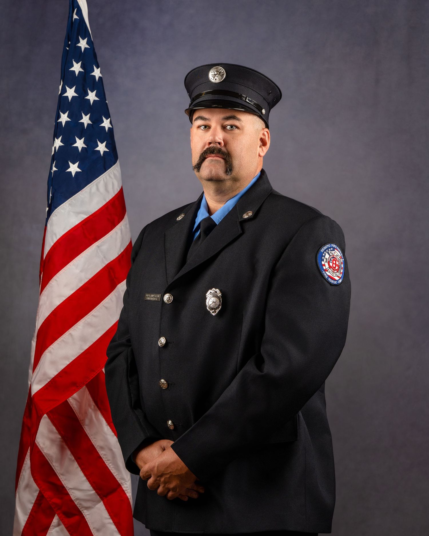 Fire department officer in dress uniform with gold insignia poses beside an American flag for an official portrait.