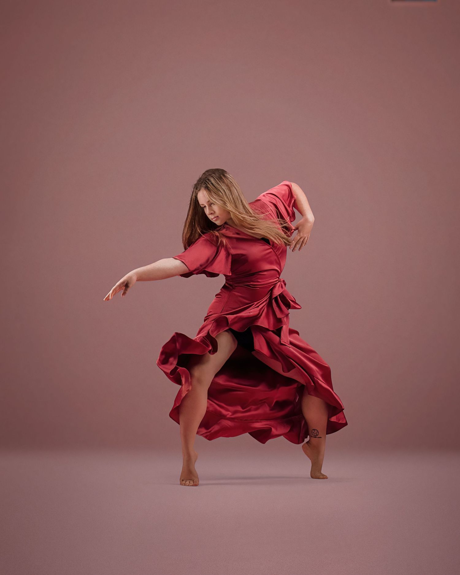 A dancer in a flowing red satin dress mid-movement against a mauve studio background.
