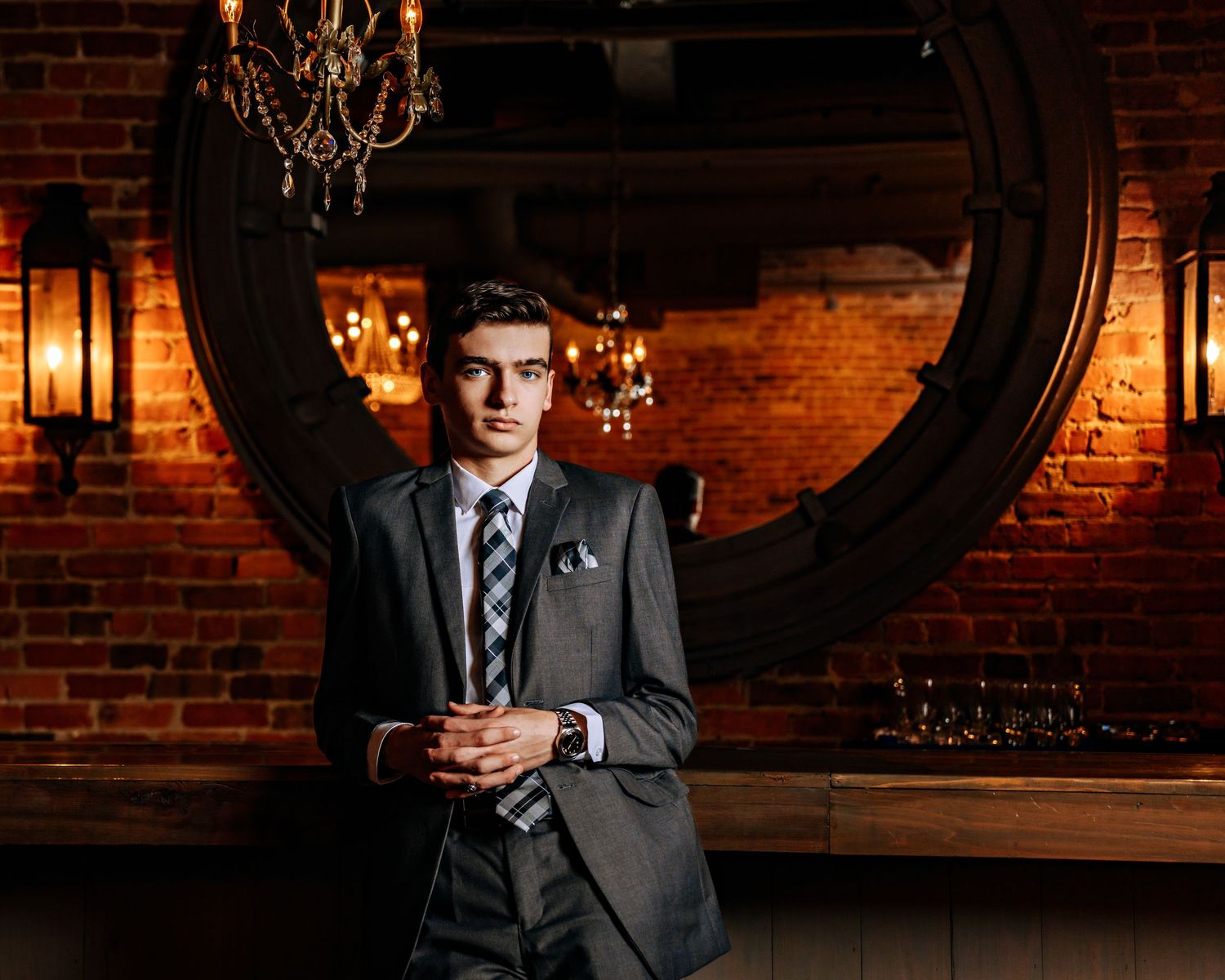 Young man in gray suit posing in rustic brick bar with chandelier.