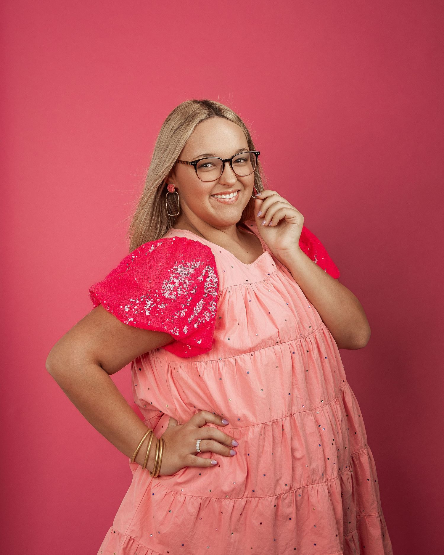 Smiling plus-size blonde woman in pink sequin dress poses against pink background.