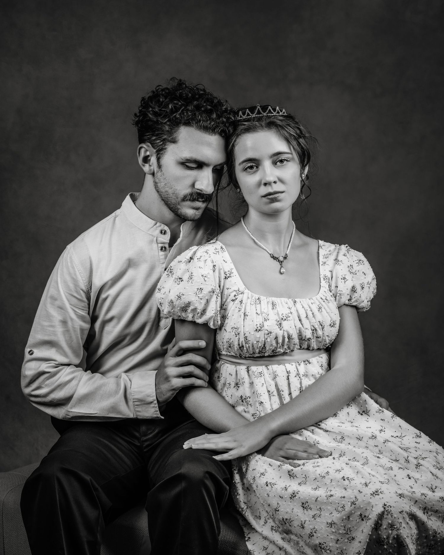A black and white portrait of a couple in period attire, the woman wearing a tiara and floral dress.