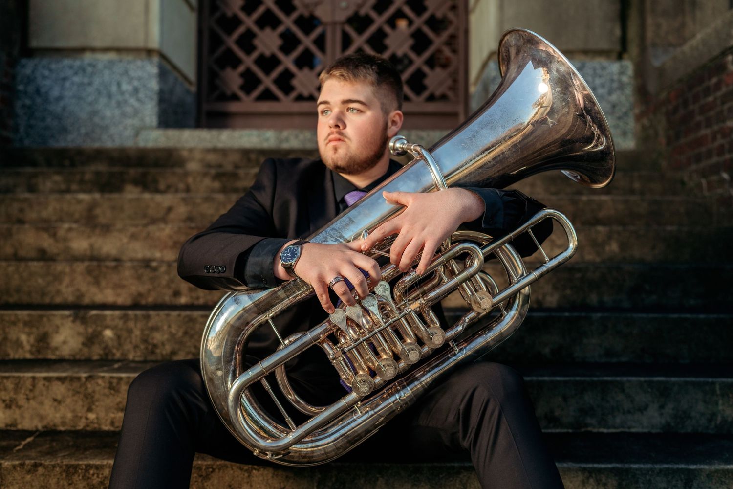 Young man in black suit sitting on steps holding a large silver tuba instrument outdoors.