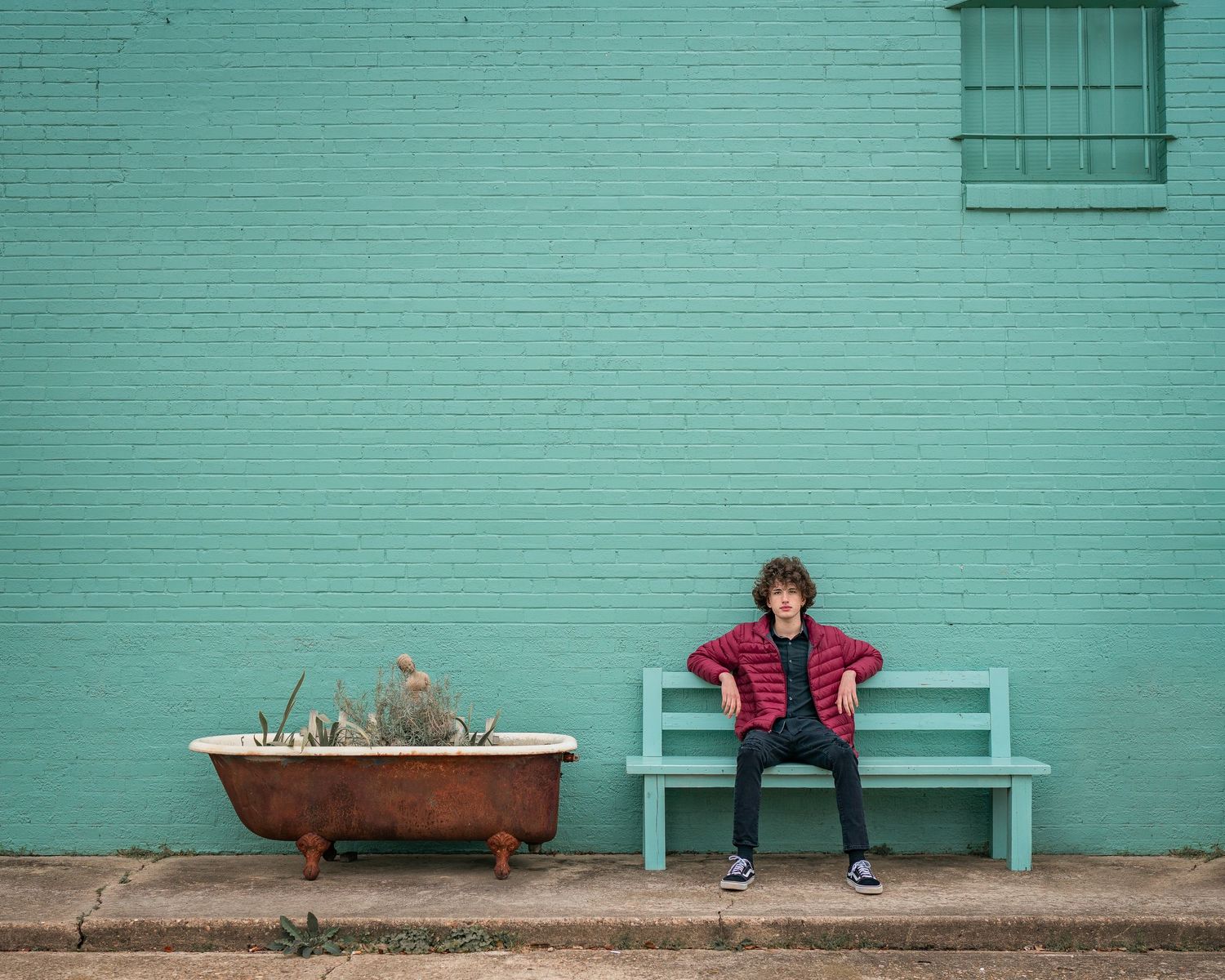 Young boy sitting on a teal bench beside a rustic bathtub planter against a green brick wall.